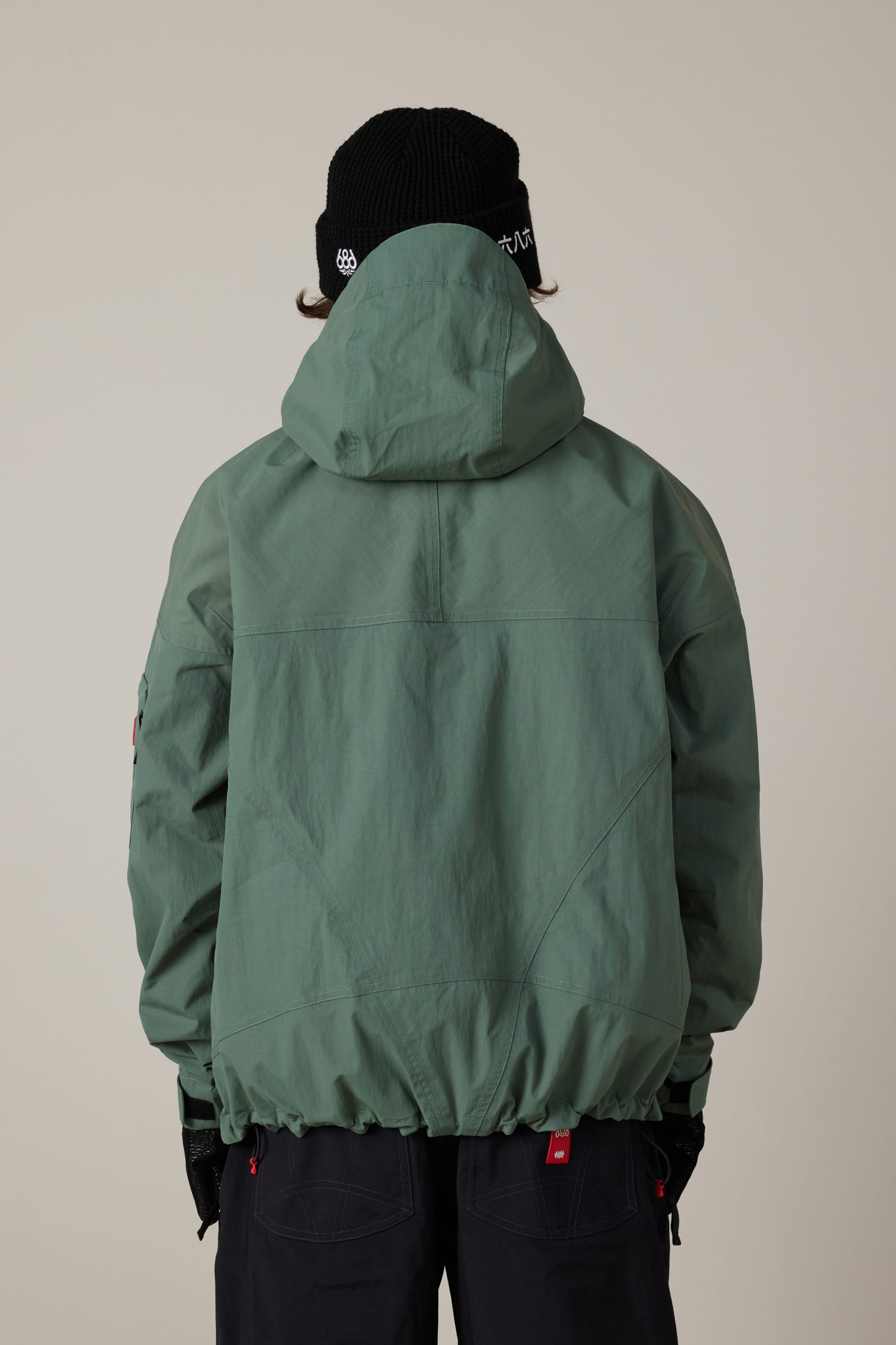 A person wearing a green 686 Men’s 686 Dojo® Shell Jacket with a hood, black gloves, black pants, and a black beanie is standing with their back to the camera against a plain light background.