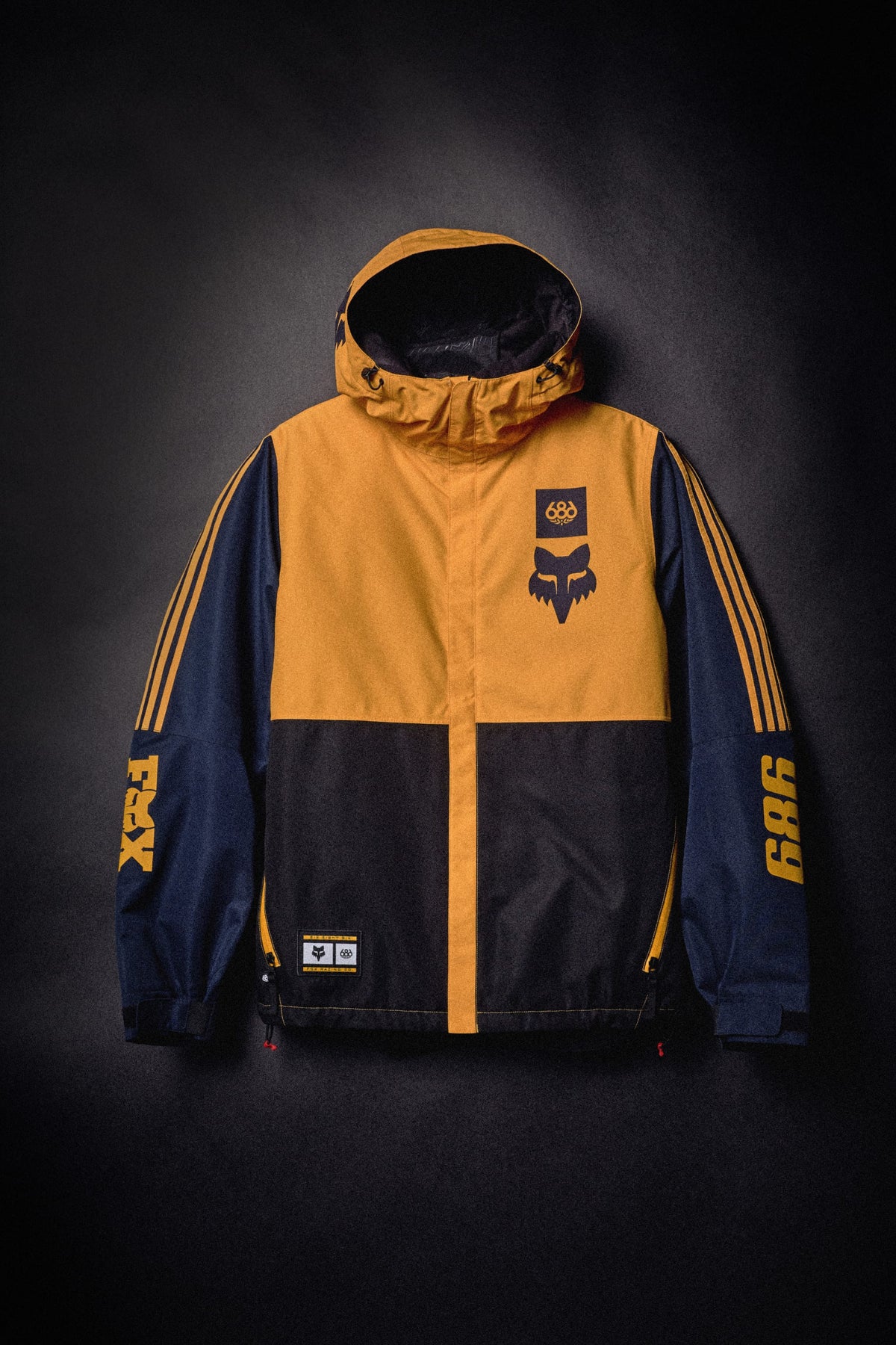 A mustard yellow and navy blue 686 Men’s Fox Racing Spec Insulated Jacket with co-branded logos on the chest and sleeves.