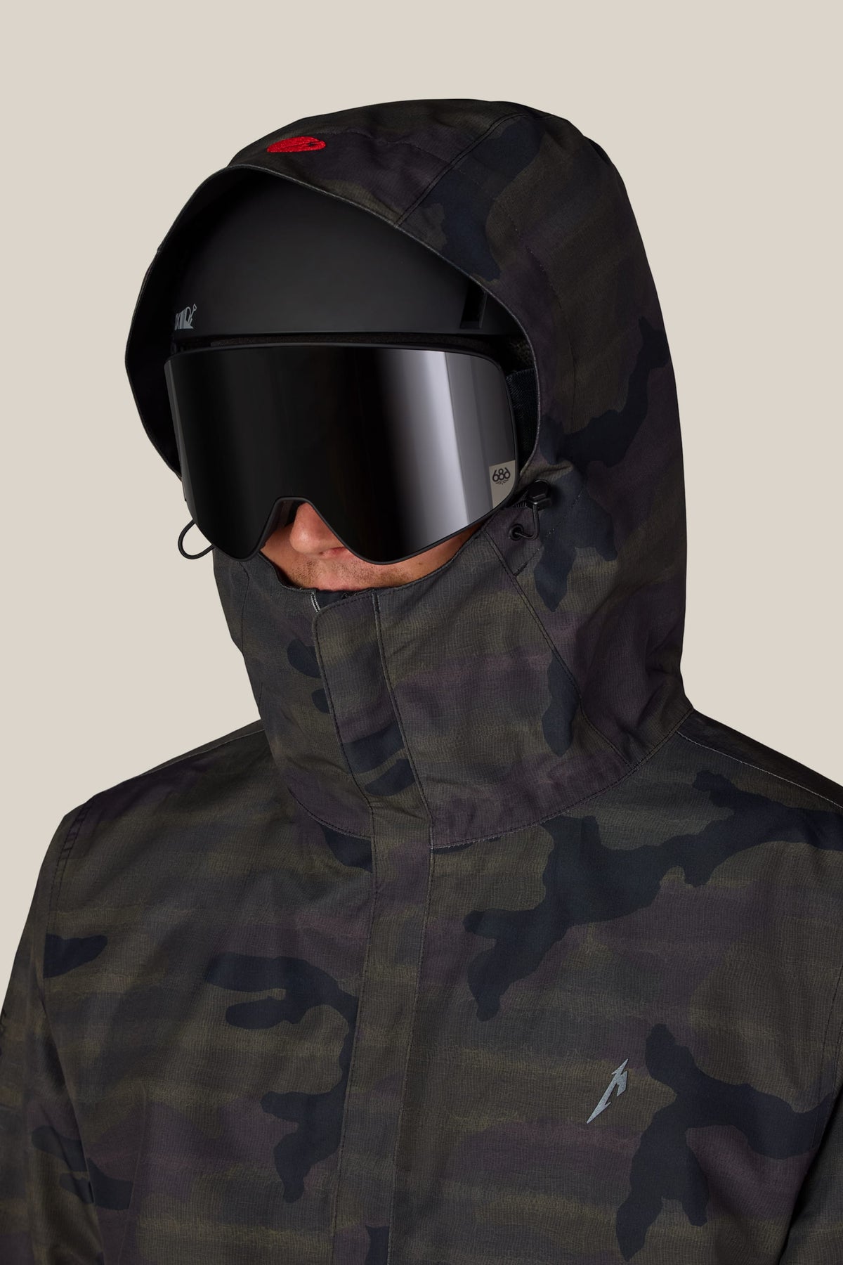 A person wearing a hooded camouflage 686 Men’s Metallica One Insulated Jacket, a black helmet, and dark ski goggles.