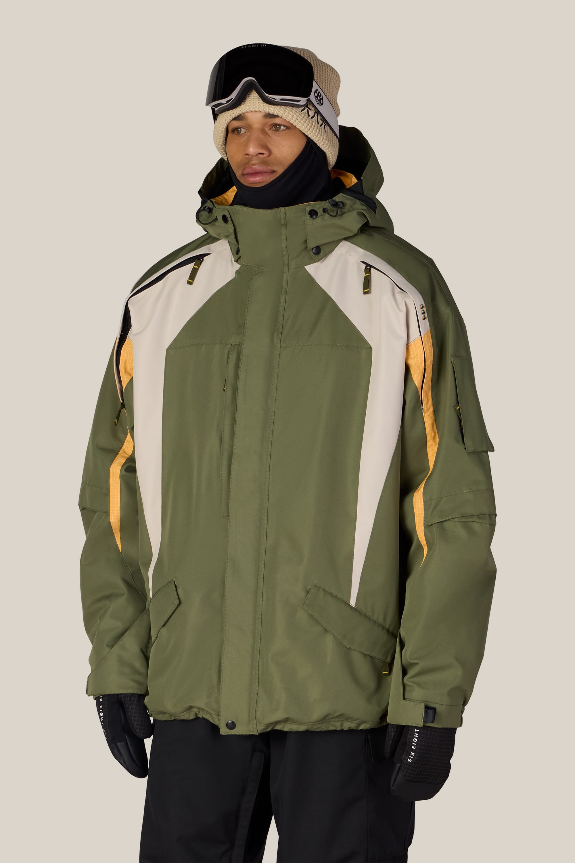 A person wearing the 686 Men’s 2003 Atom Jacket in green and beige with yellow accents, paired with black ski gloves, ski goggles over a beige beanie, and a black neck gaiter, stands against a plain light background.