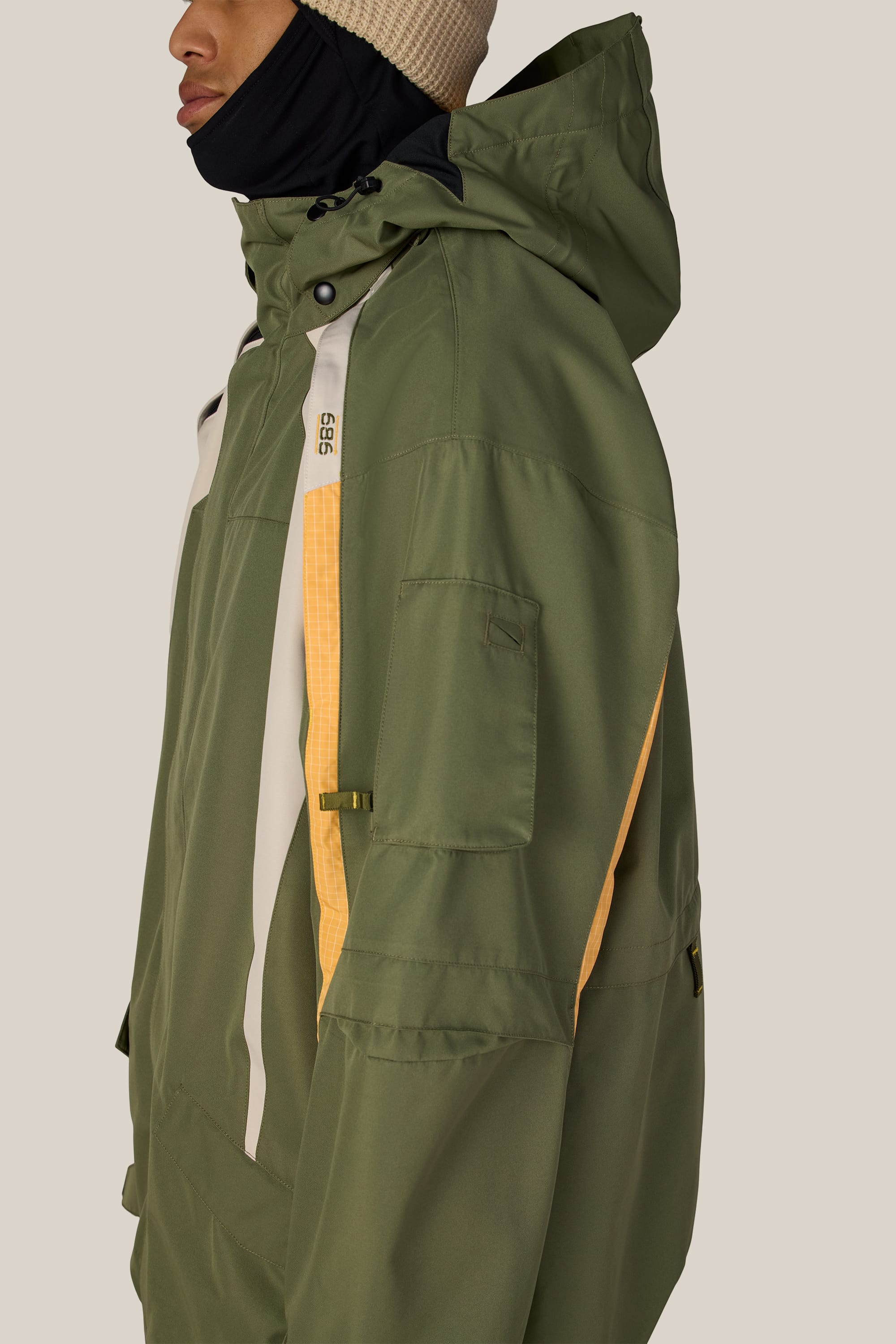 A person wearing the 686 Men’s 2003 Atom Jacket in olive green with yellow and white accents, paired with a beige knit beanie and a black face covering, is shown from the side against a plain background.
