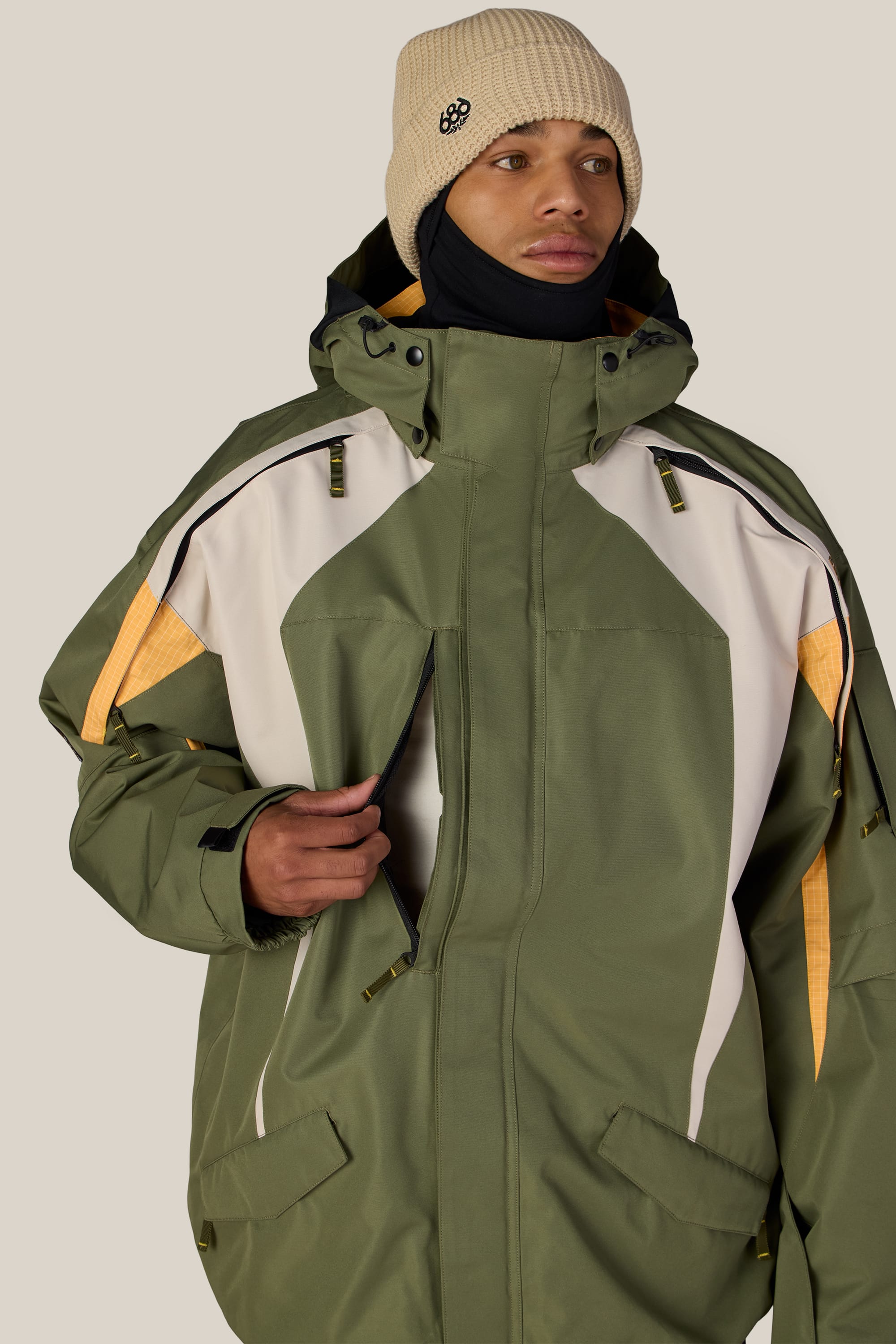 A person wearing the 686 Men’s 2003 Atom Jacket—a green and white waterproof jacket with yellow accents—along with a beige knit beanie and a black neck warmer, standing against a plain light background.