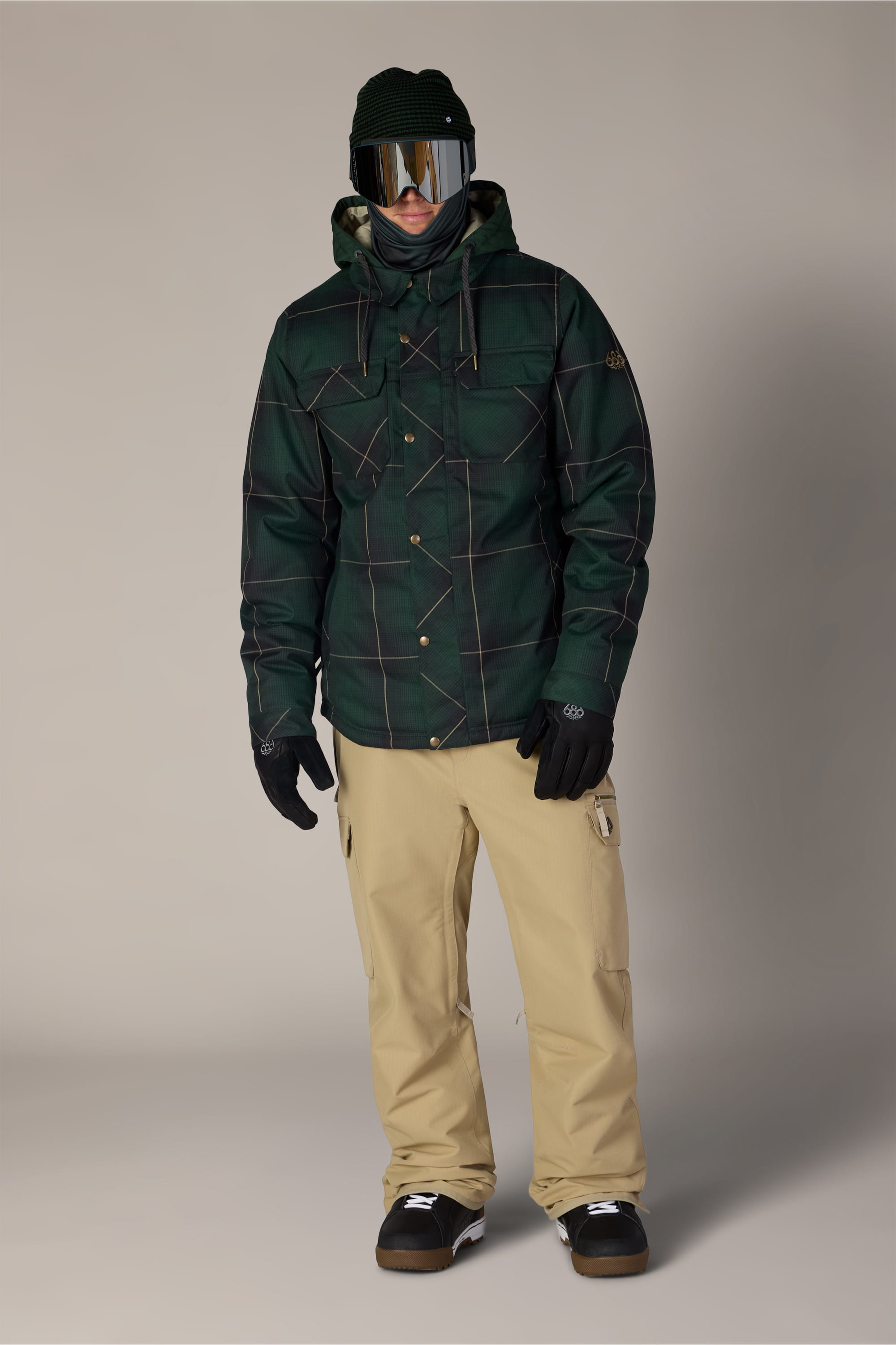A person wearing a green plaid jacket, 686 Men’s 686 Nightline Cargo Pant in tan, black gloves, black beanie, and reflective ski goggles stands against a plain background.