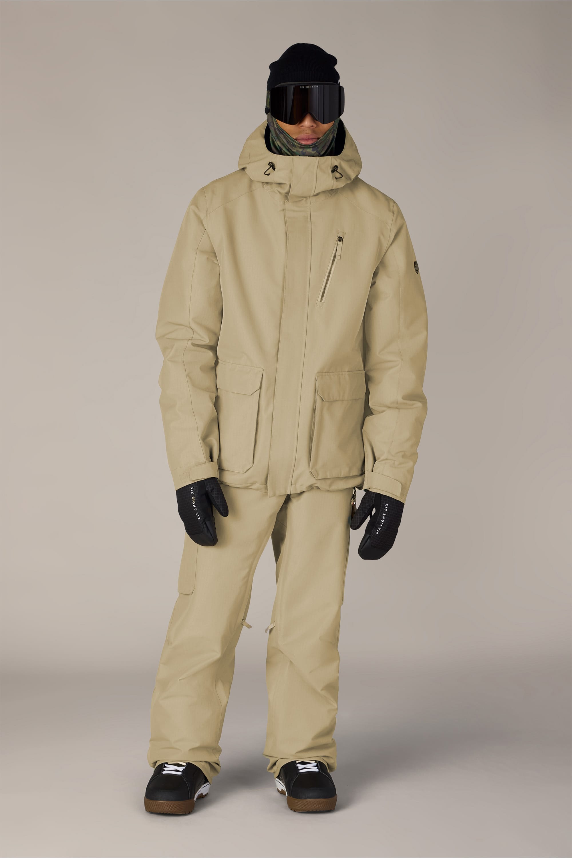 A person wearing the 686 Men’s Nightline Cargo Pant—a pair of men’s woven pants by 686 crafted from waterproof polyester fabric—paired with a beige ski jacket, black gloves, ski goggles, a black beanie, and dark boots, stands facing forward against a plain background.