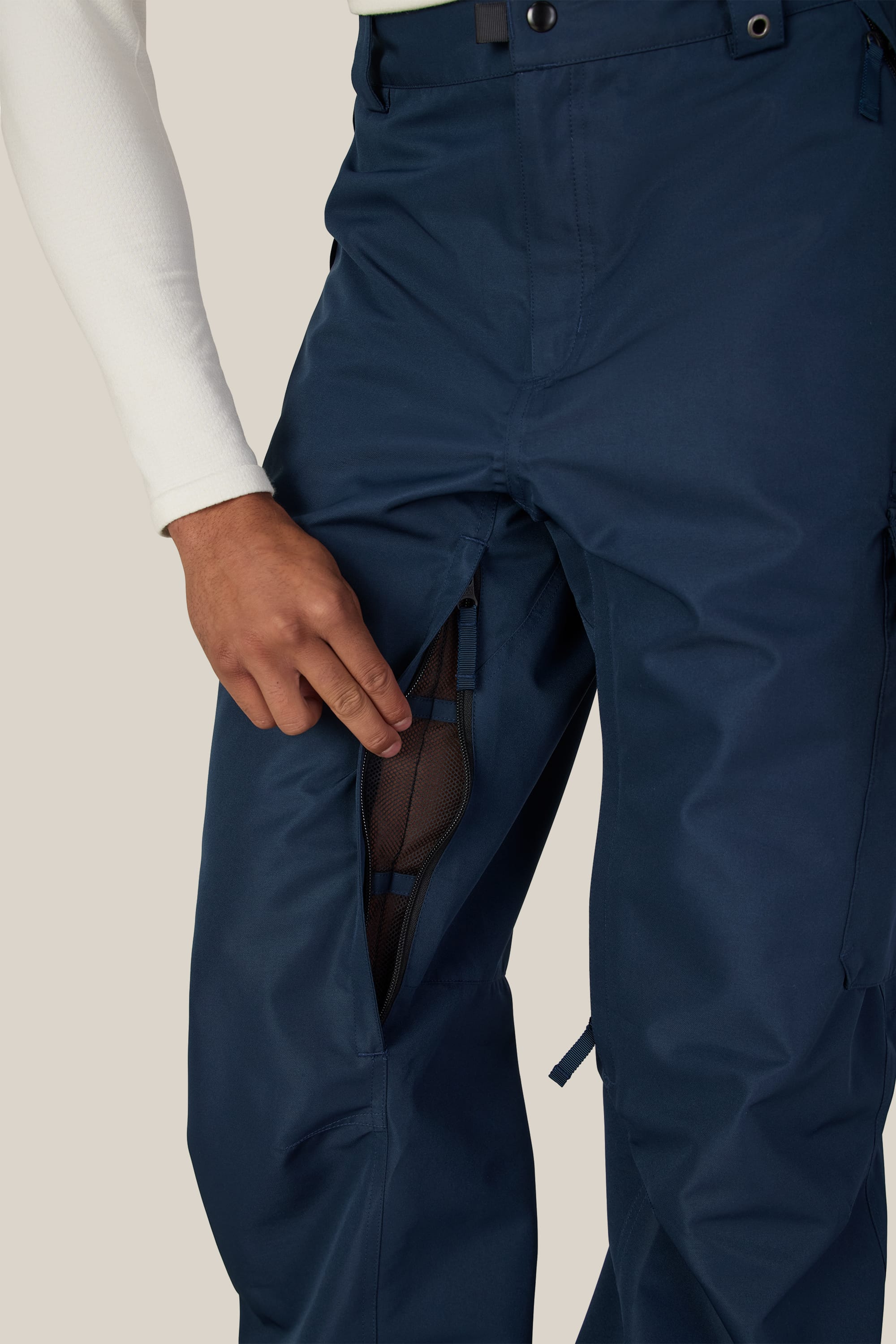 A person wearing the 686 Men’s 686 Nightline Cargo Pant in navy blue, featuring a zippered mesh vent on the thigh that reveals a black mesh lining. The waterproof polyester pants by 686 are paired with a long-sleeve white shirt.