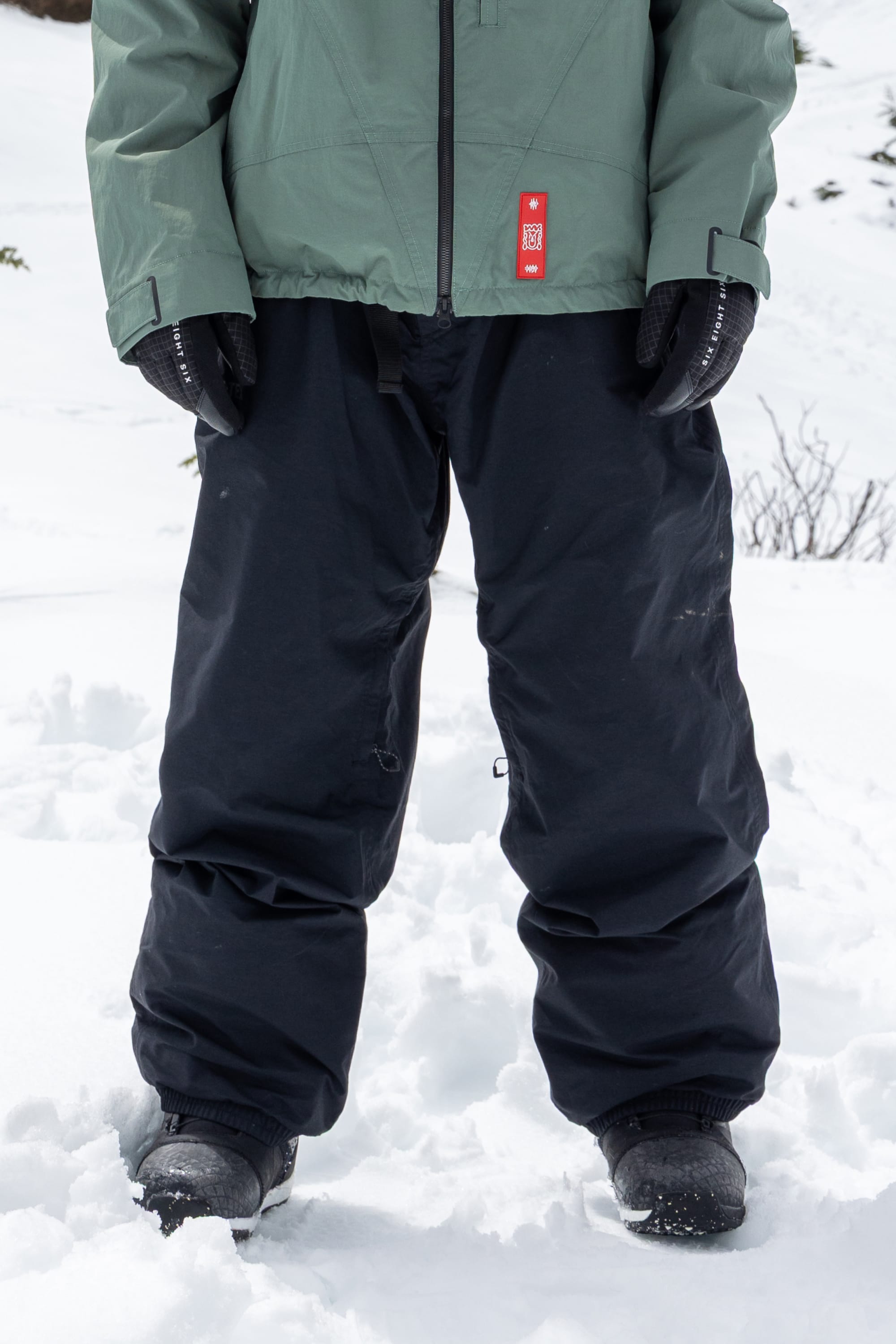 A person stands in the snow, with only their lower half visible, wearing 686 Men’s 686 Dojo® GORE-TEX® Shell Pants surrounded by snow-covered ground.