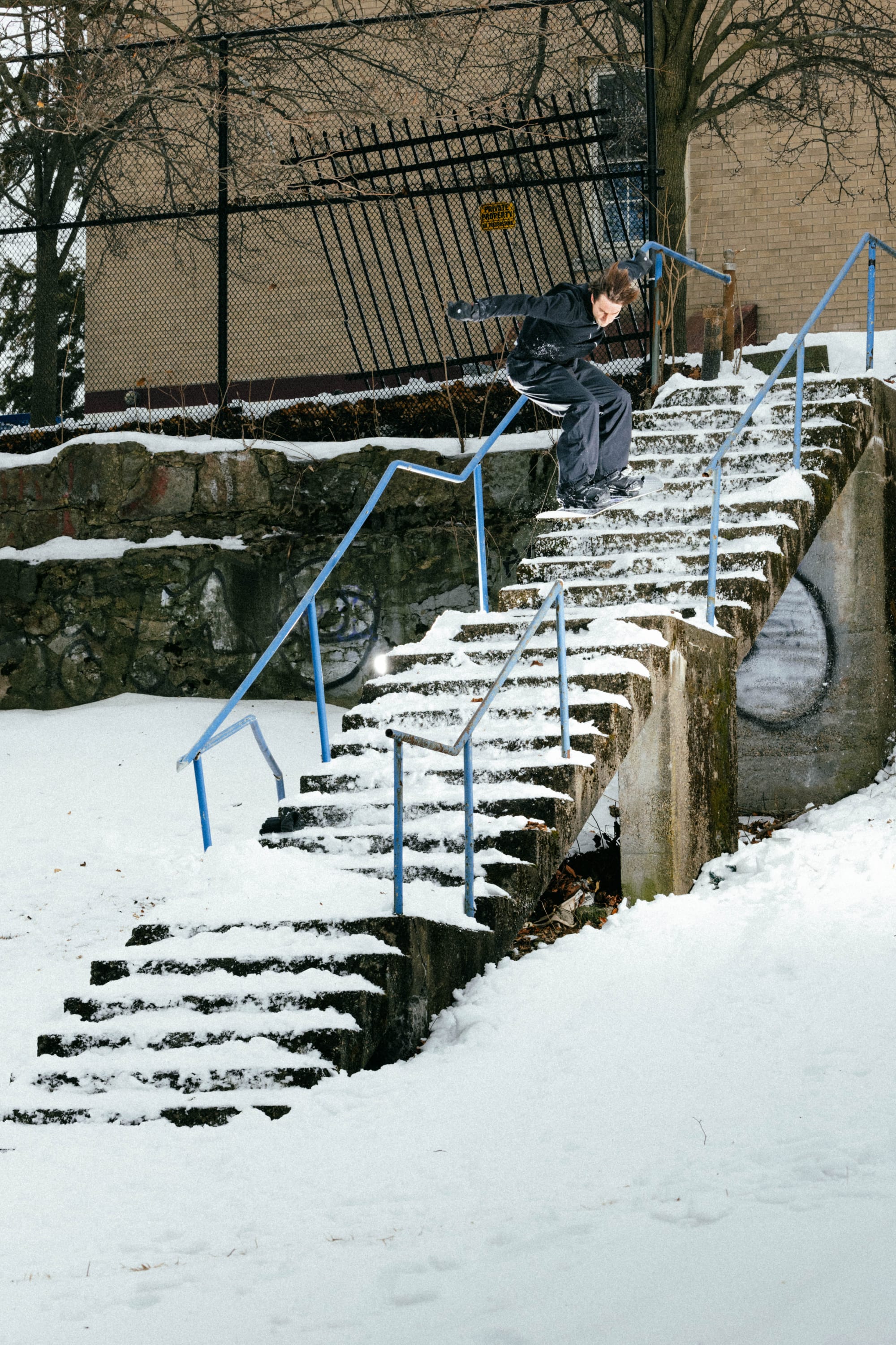 Wearing the 686 Men’s 686 Dojo® GORE-TEX® Shell Pant from 686, a person skateboards down a snow-covered handrail on an outdoor stairway, surrounded by snowy ground and bare trees, with a metal fence and brick building in the background.