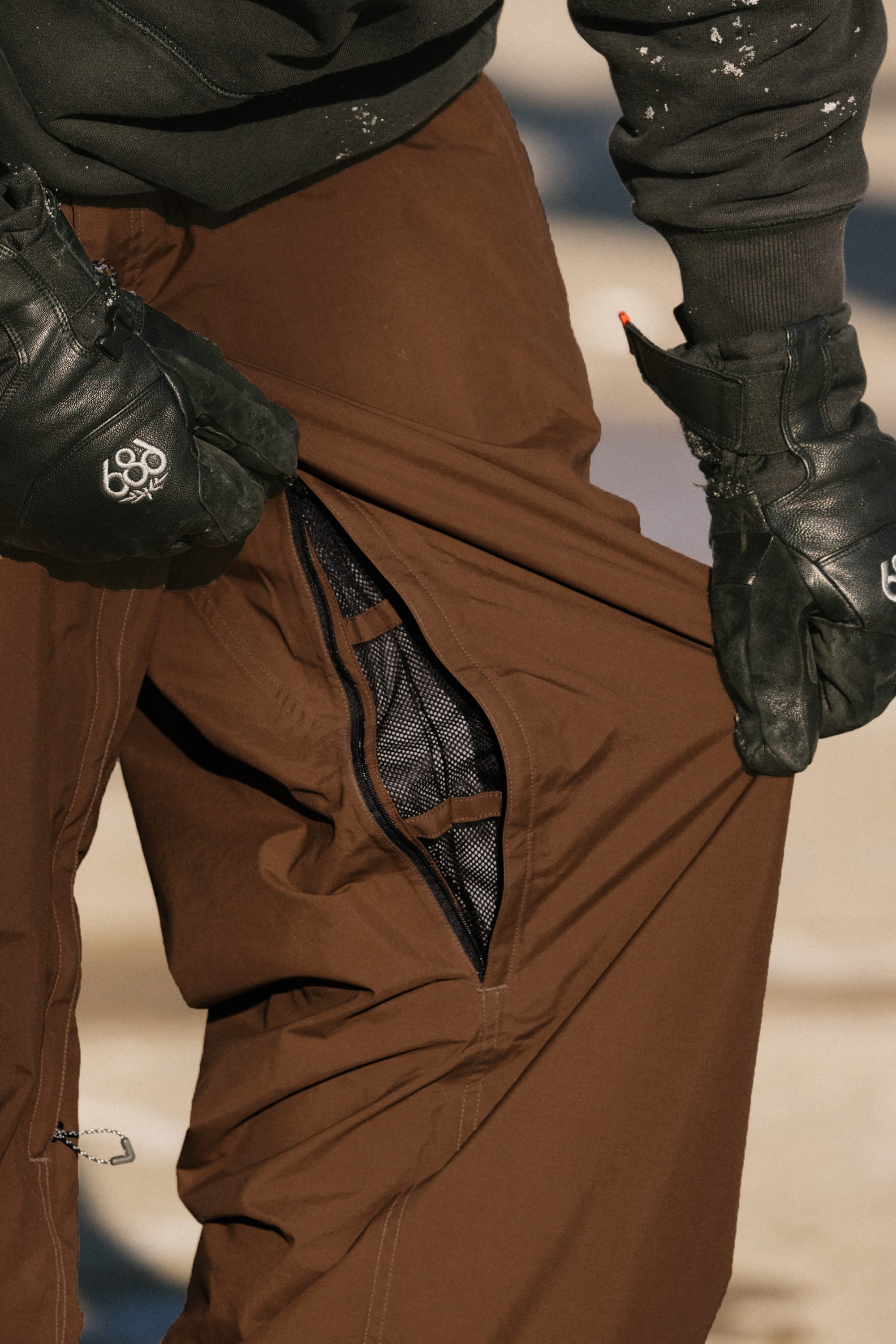A person wearing black gloves pulls open a mesh-lined ventilation zipper on their brown 686 Men’s 686 Dojo® GORE-TEX® Shell Pant, revealing the inner material of these waterproof pants by 686.