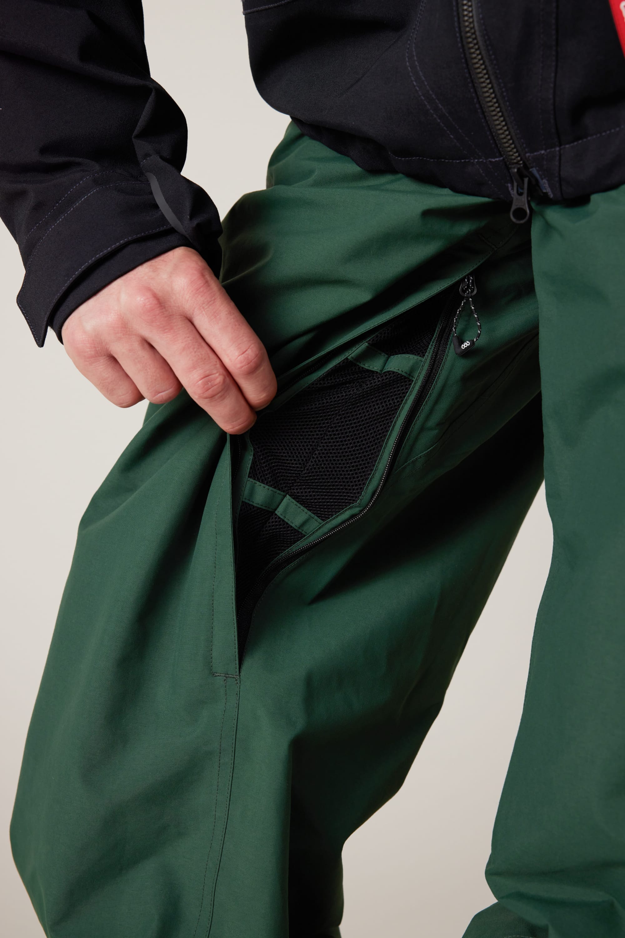 A person wearing 686 Men’s Dojo® GORE-TEX® Shell Pants in green unzips a mesh-lined pocket to reveal the inside. The person is also wearing a black jacket.