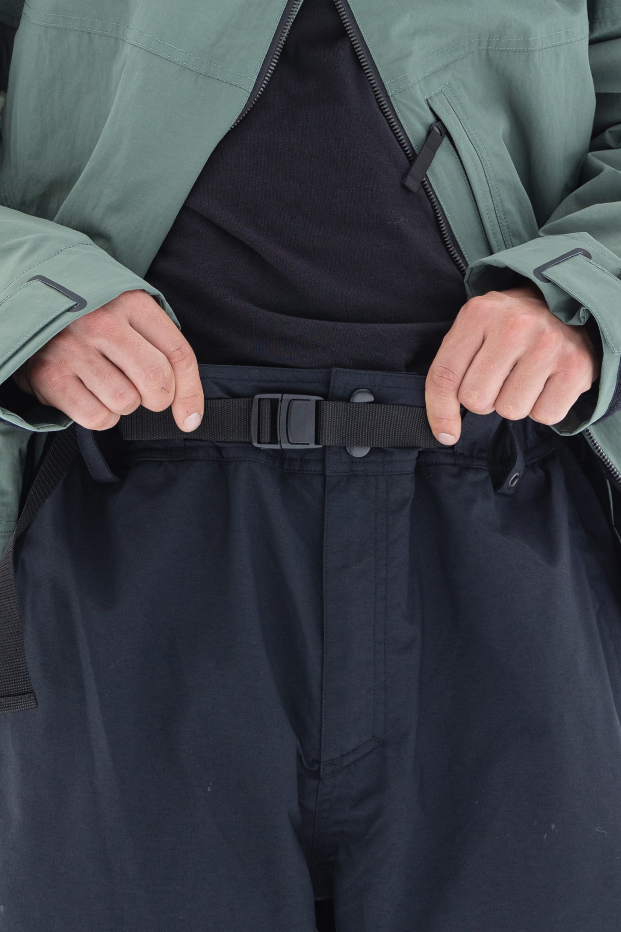 A person adjusts a black webbing belt on the 686 Men’s 686 Dojo® Shell Pant in navy blue, paired with a black shirt and a green jacket. Only the torso and hands are visible.
