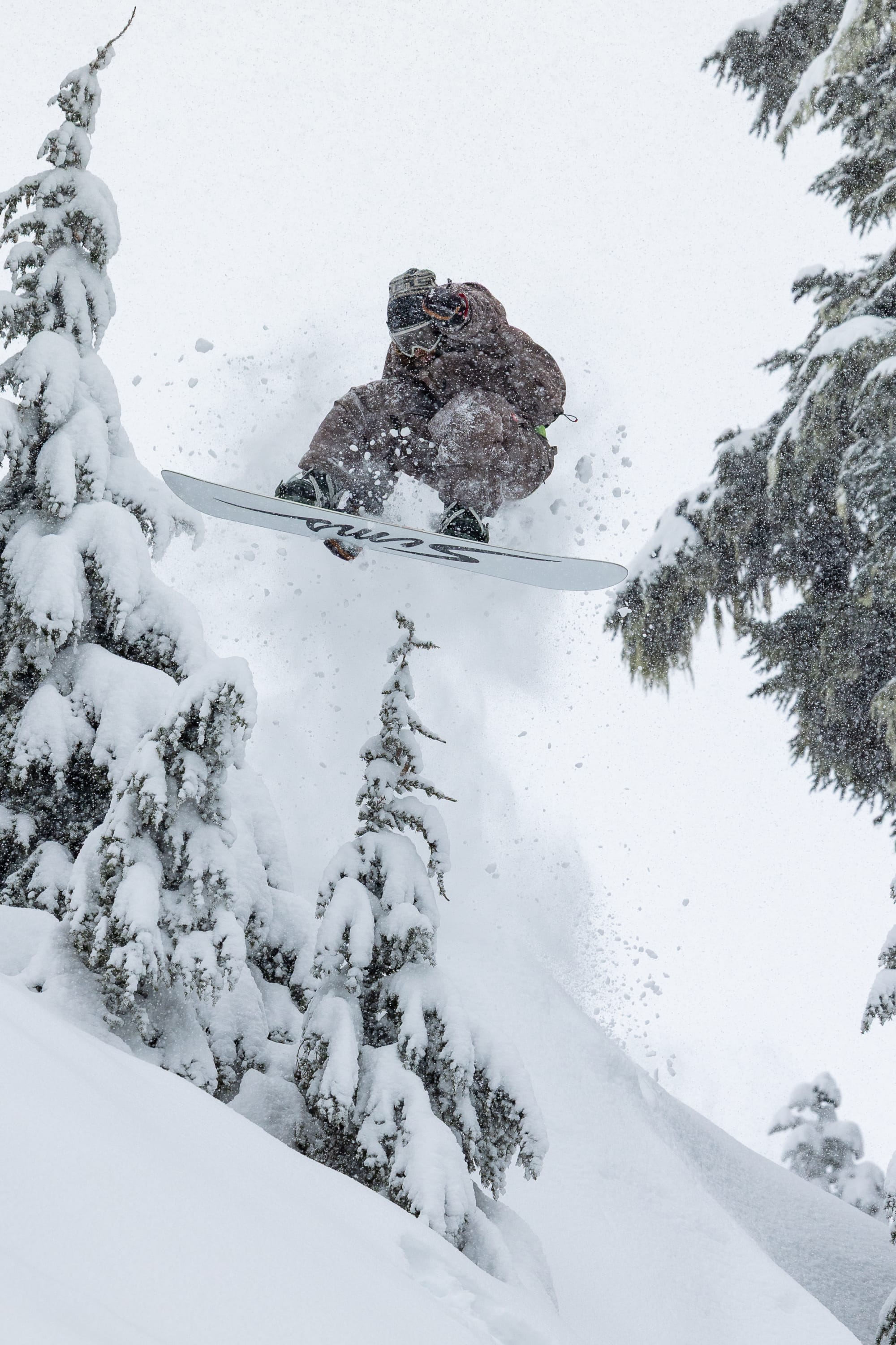 A snowboarder in mid-air, wearing the 686 Men’s 686 Dojo® Shell Pant by 686, leaps off a snowy slope surrounded by snow-covered pine trees on a wintery mountain, with snow spraying behind them as they descend.