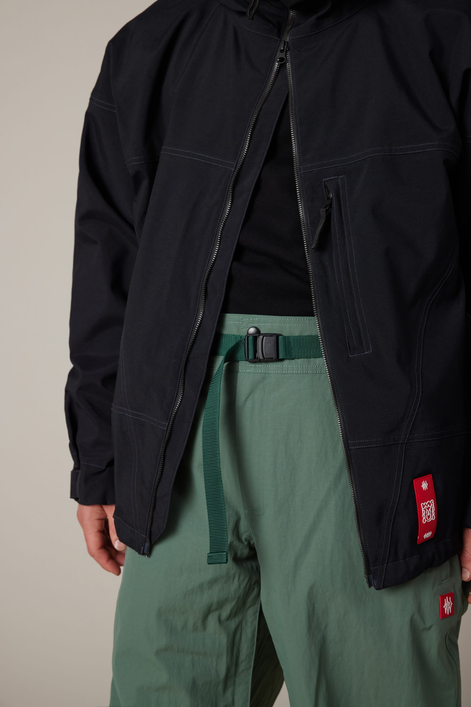 A person wearing a black jacket, black shirt, green belt, and light green 686 Men’s Dojo® Shell Pant by 686, featuring a red patch. The jacket is partially unzipped. The background is plain and neutral.