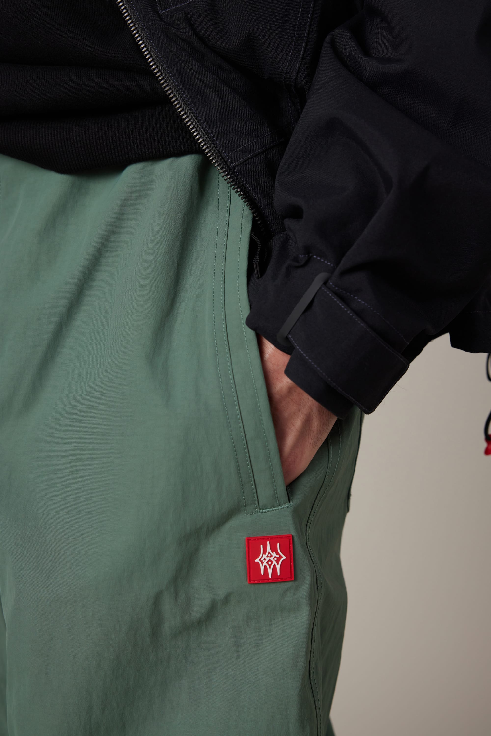 A person wearing light green 686 Men’s 686 Dojo® Shell Pants with a red patch and a black jacket has their hand in their pocket.