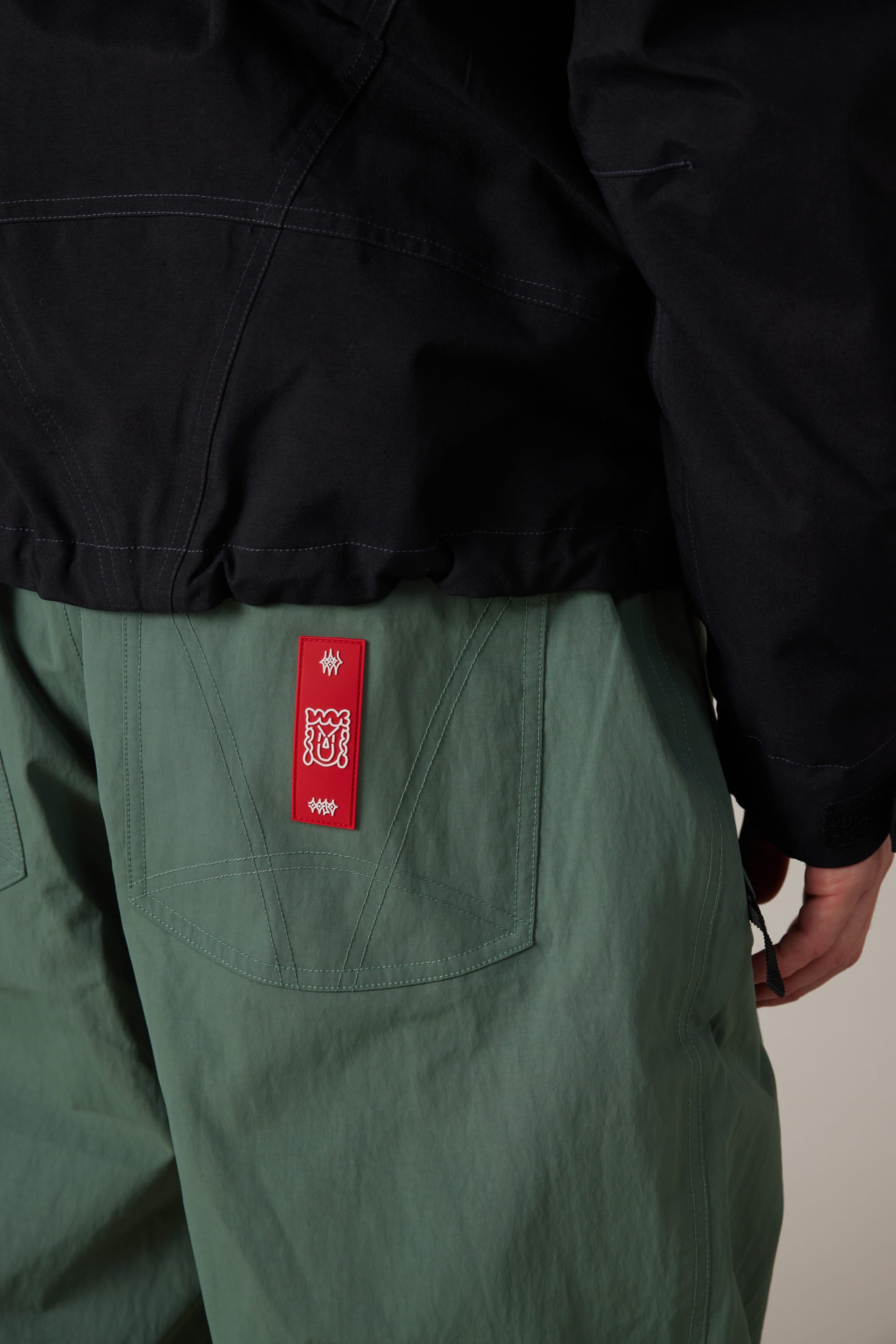 A person wearing 686 Men’s 686 Dojo® Shell Pant in green, featuring a visible red tag above the back pocket, paired with a dark jacket; shown from the waist down and facing away from the camera.