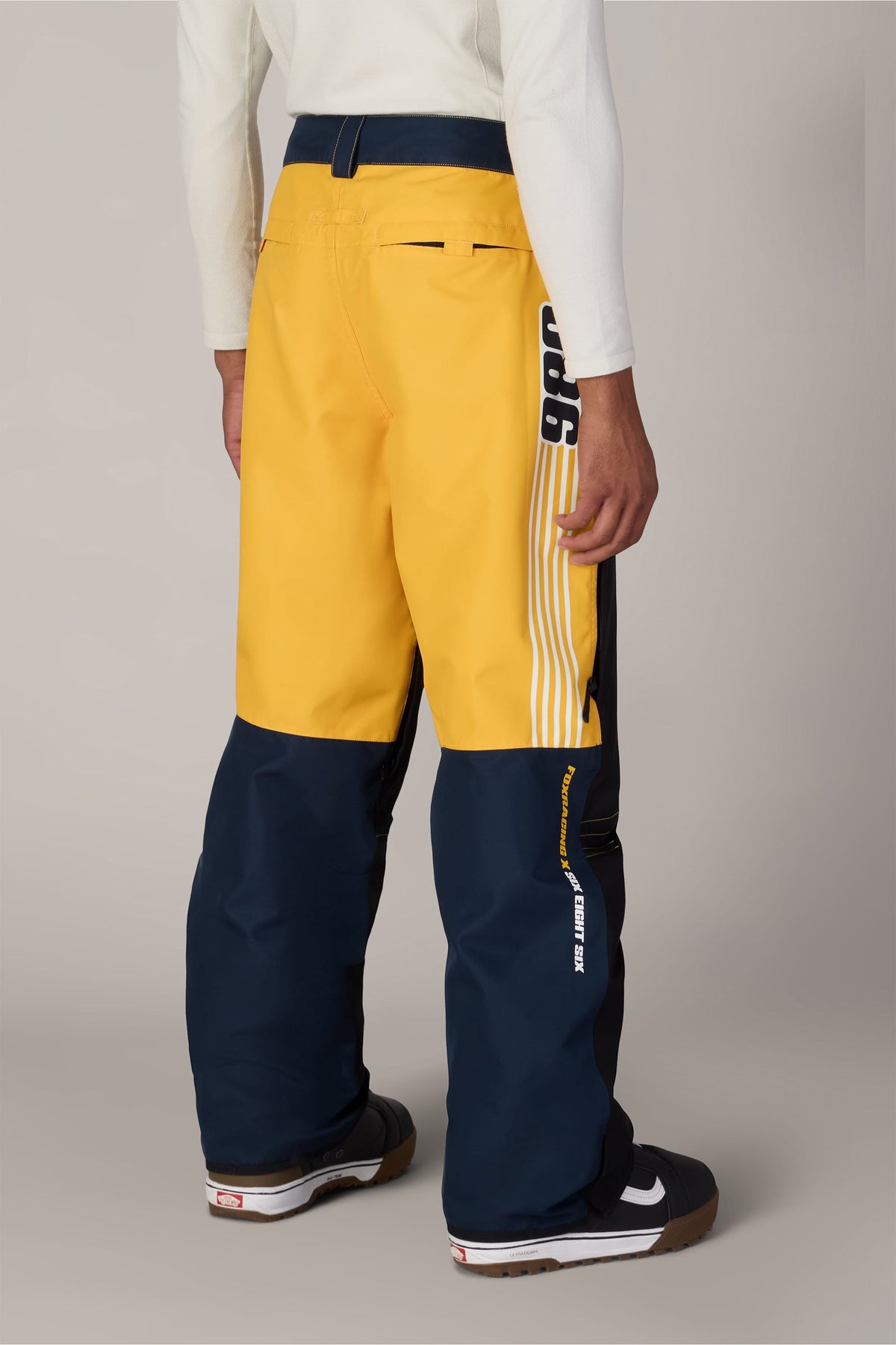 A person wearing yellow and navy blue 686 Men’s Fox Racing Spec Shell Pants with side racing stripes and branding.
