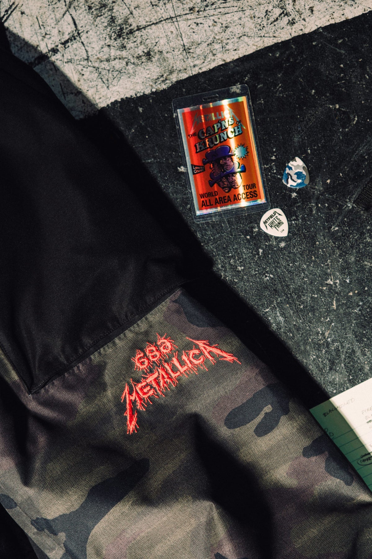 686 Men’s Metallica One Shell Pant in camouflage with a red embroidered Metallica logo, displayed with a Metallica backstage pass, two guitar picks, and a notebook on a dark surface.