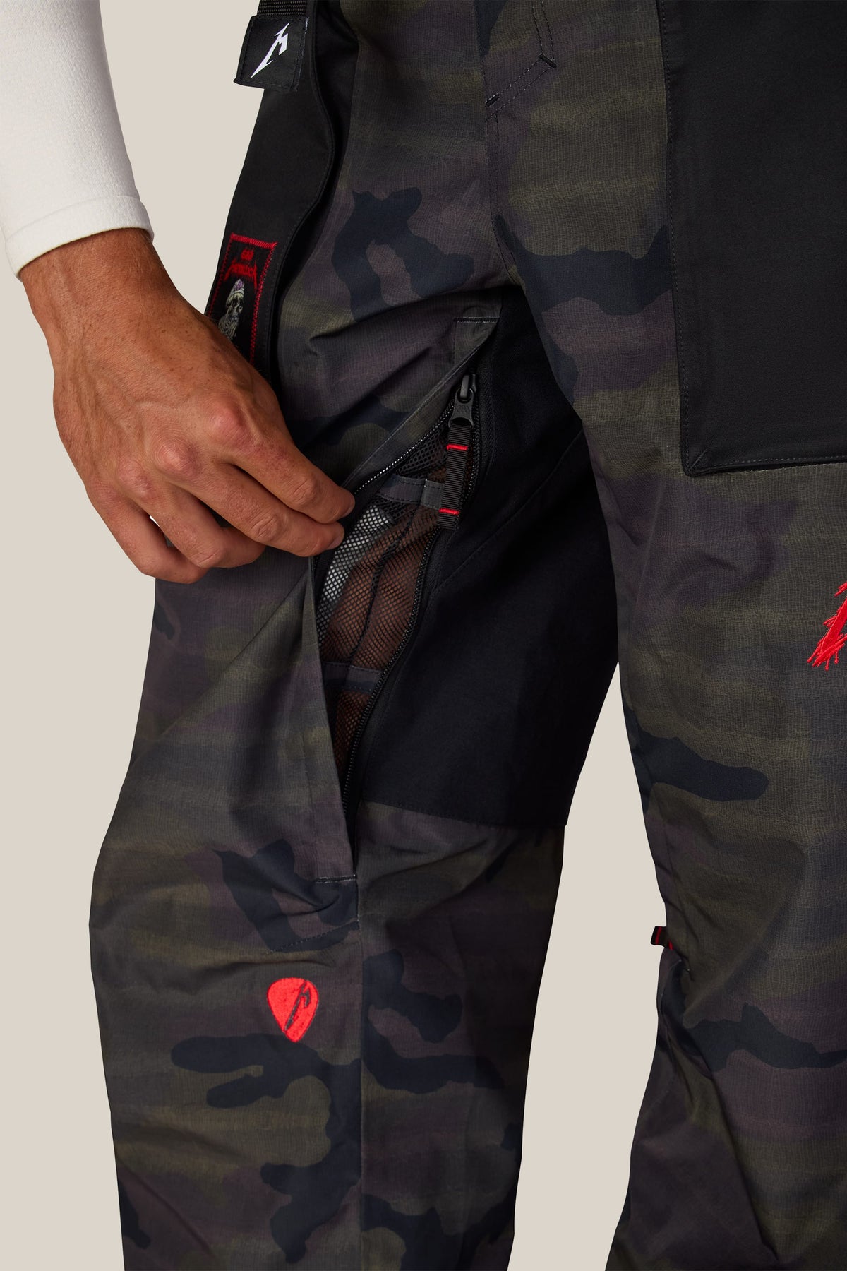 A hand opens a mesh-lined zippered pocket on the thigh of 686 Men’s Metallica One Shell Pant dark camouflage waterproof pants.