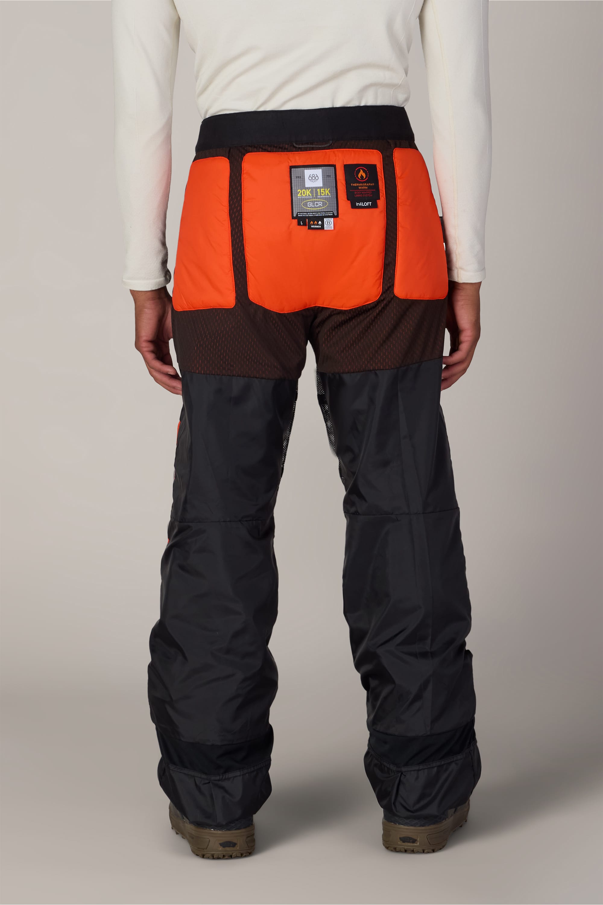 A person faces away, wearing a white long-sleeve shirt and the 686 Men’s Hydra™ Thermagraph® Pant by 686—black and orange waterproof woven pants featuring mesh sections and a label on the back—standing on a neutral background.