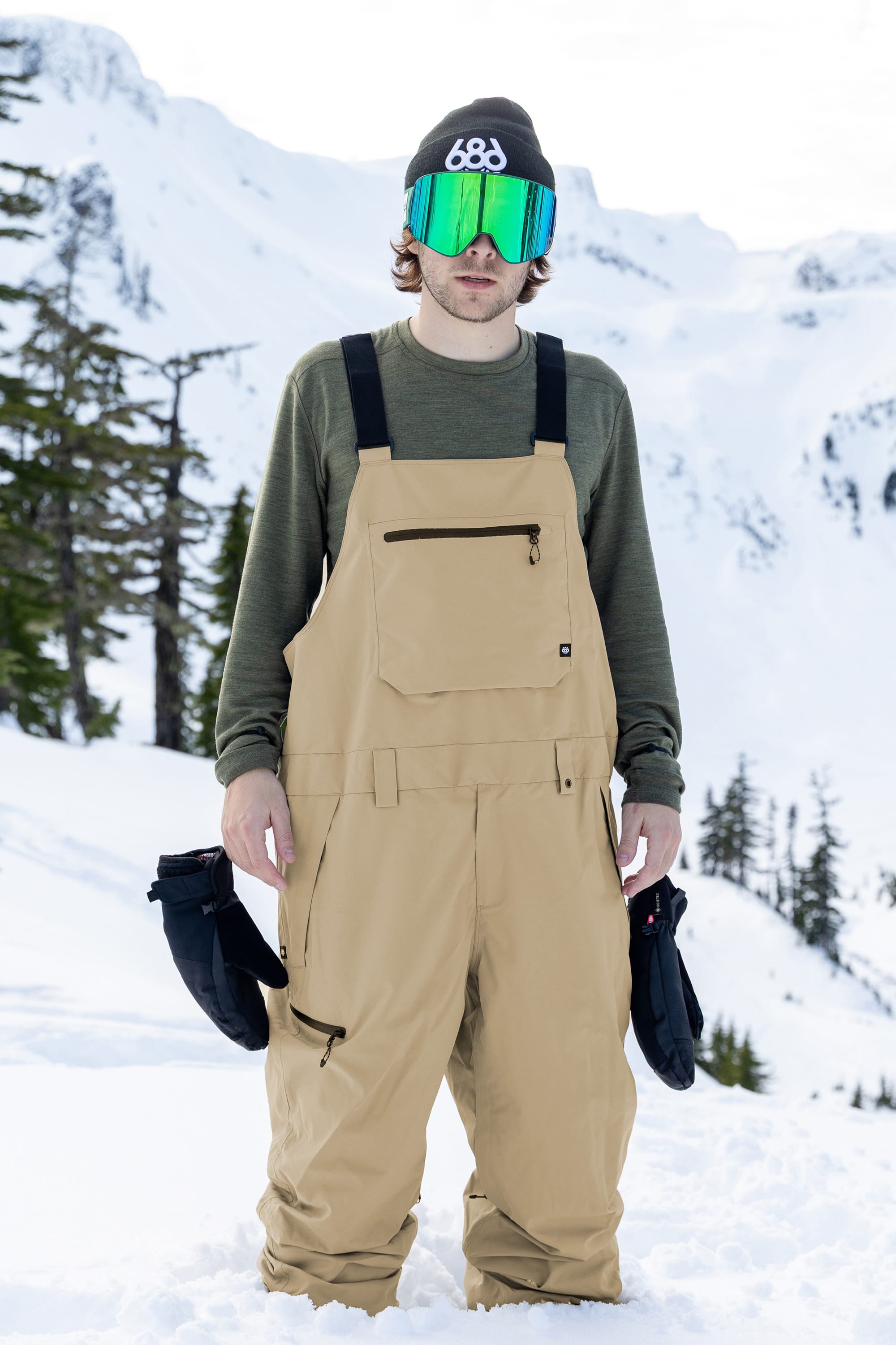 Wearing the 686 Men’s Lockup™ Shell Bib in tan, along with a green sweatshirt, mirrored goggles, and a beanie, a person stands in snowy mountains holding black gloves.