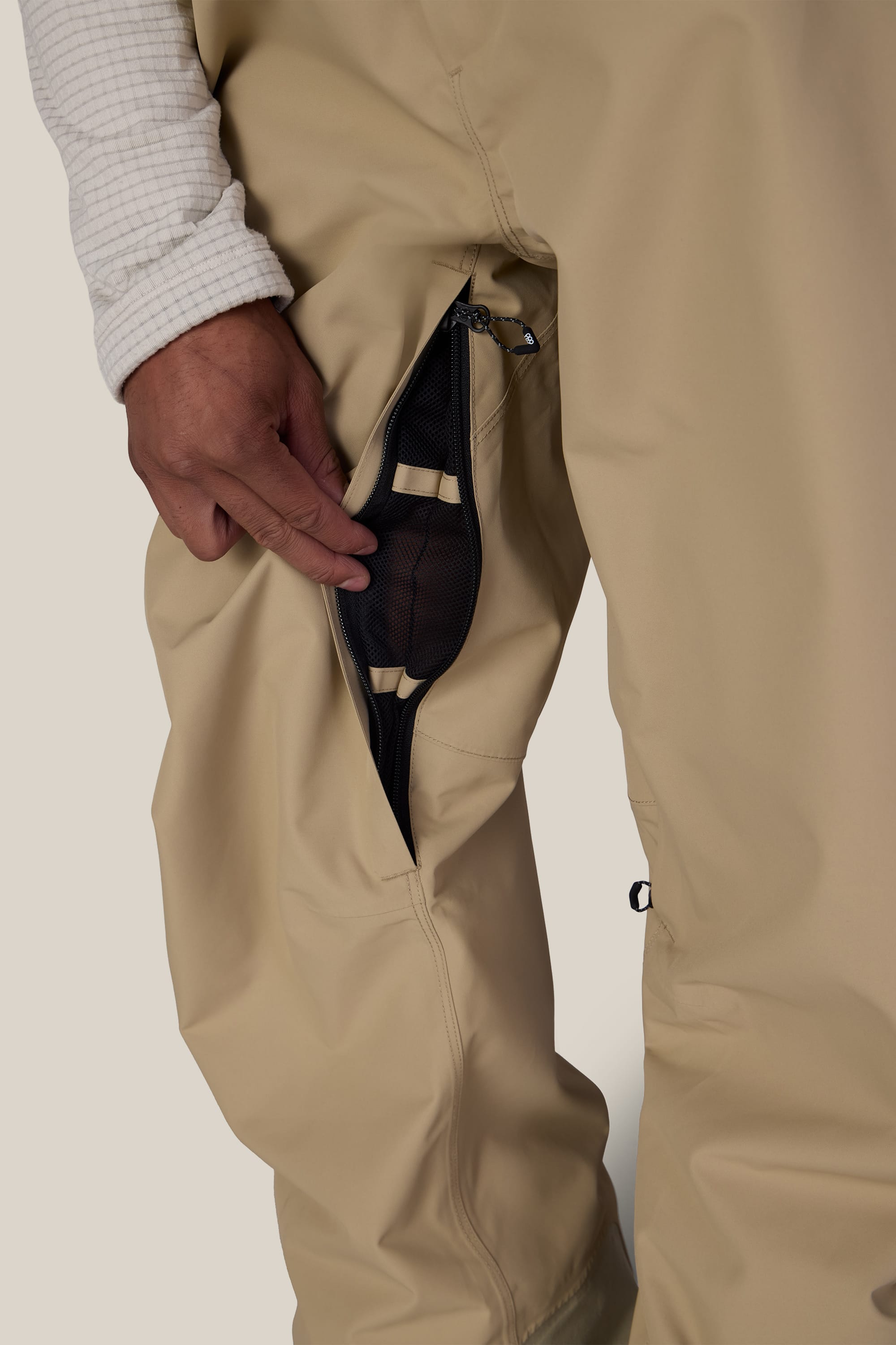 A person wearing the 686 Men’s Lockup™ Shell Bib in beige opens its zippered side pocket to reveal a black mesh lining inside. The person’s hand and part of a white, textured shirt sleeve are visible.