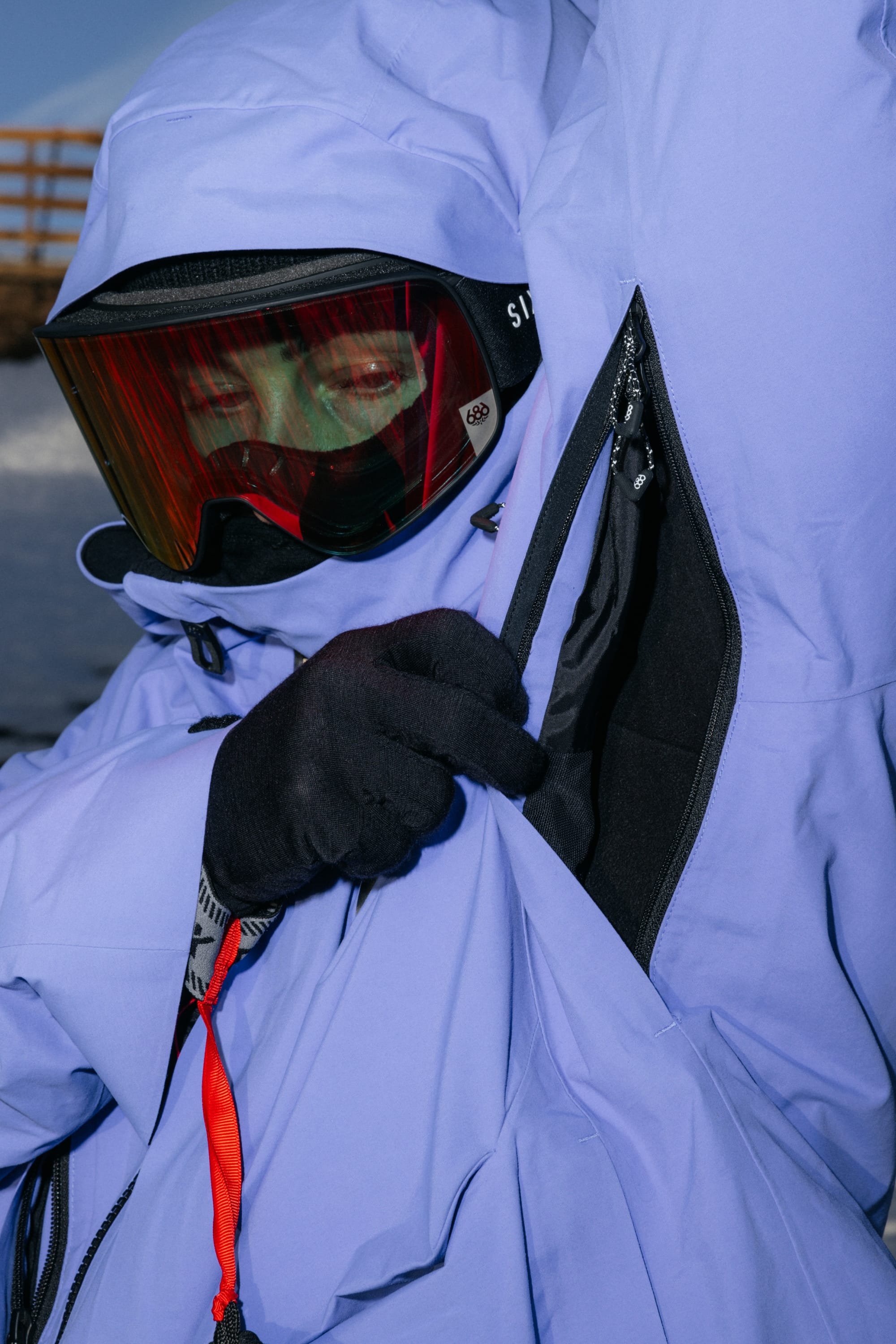 Wearing the 686 Women’s GORE-TEX® Scheme Thermagraph® Jacket, a person zips up their blue jacket outdoors on a snowy day, paired with black gloves and red ski goggles.