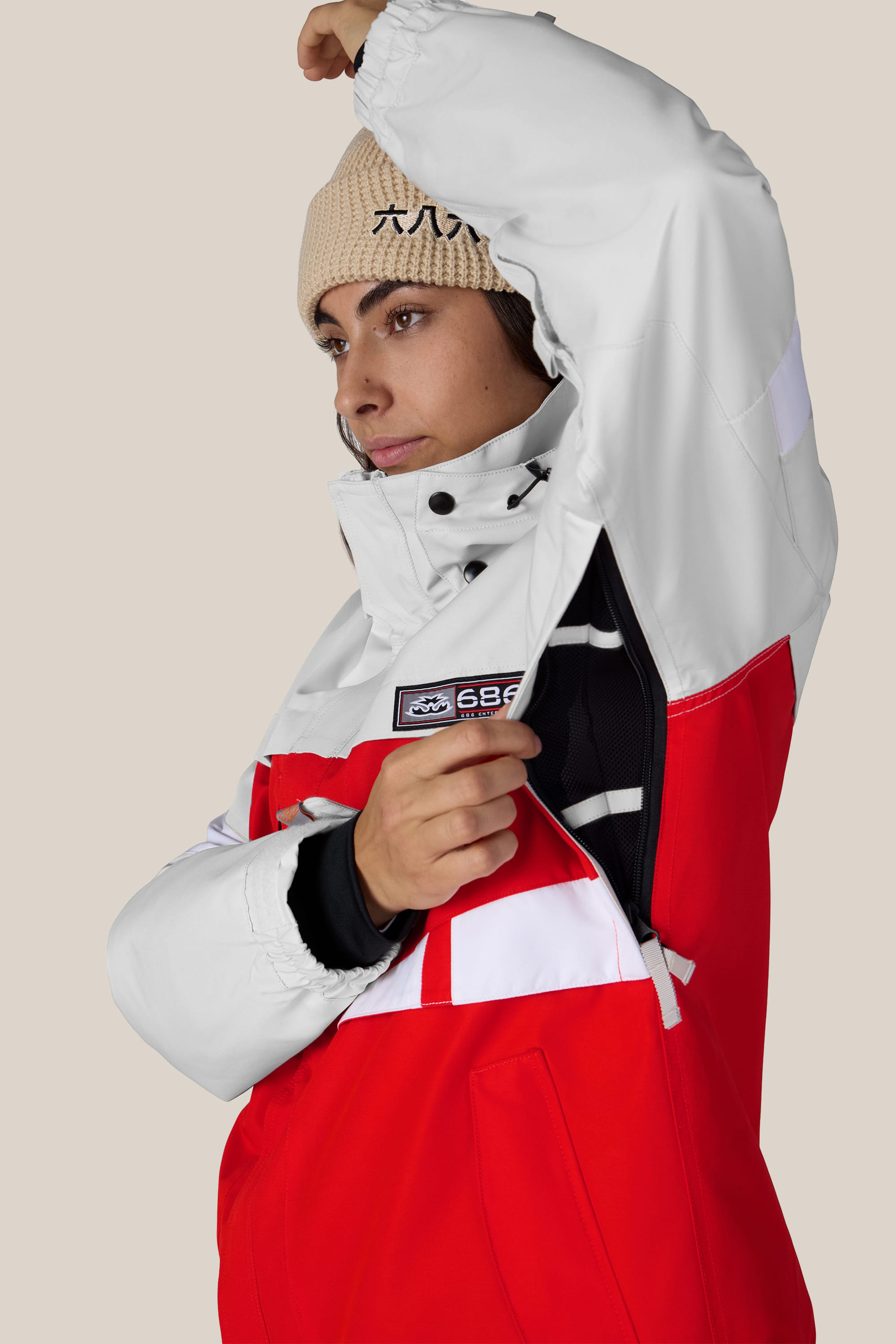Wearing the 686 Women’s Search Jacket in red and white, a person unzips an underarm vent. They pair the waterproof jacket with a beige beanie, looking to the side against a plain background.