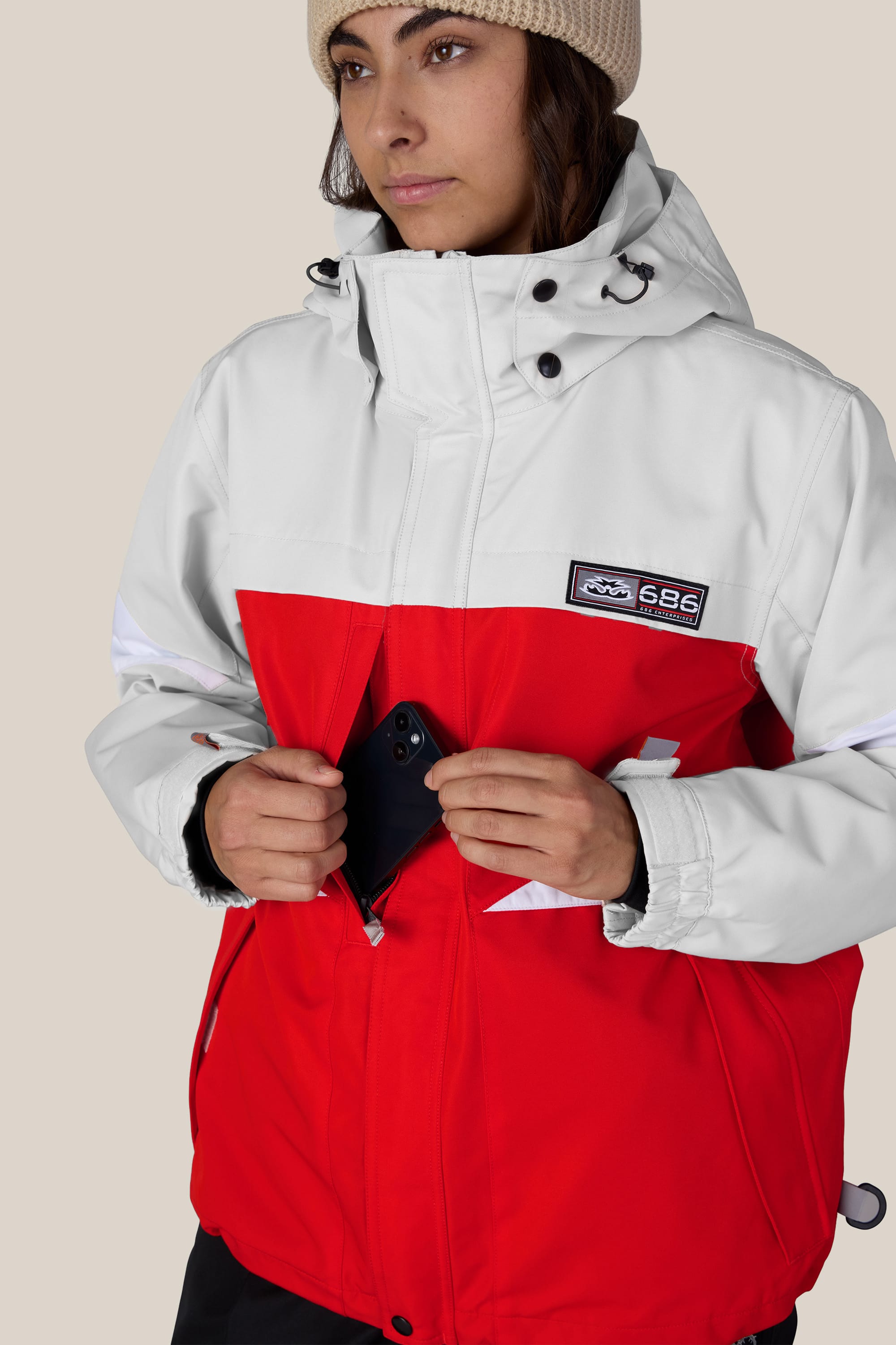 Wearing the 686 Women’s Search Jacket by 686, a person in a red and white woven jacket and beige beanie places a black smartphone into the front zippered pocket of their jacket.
