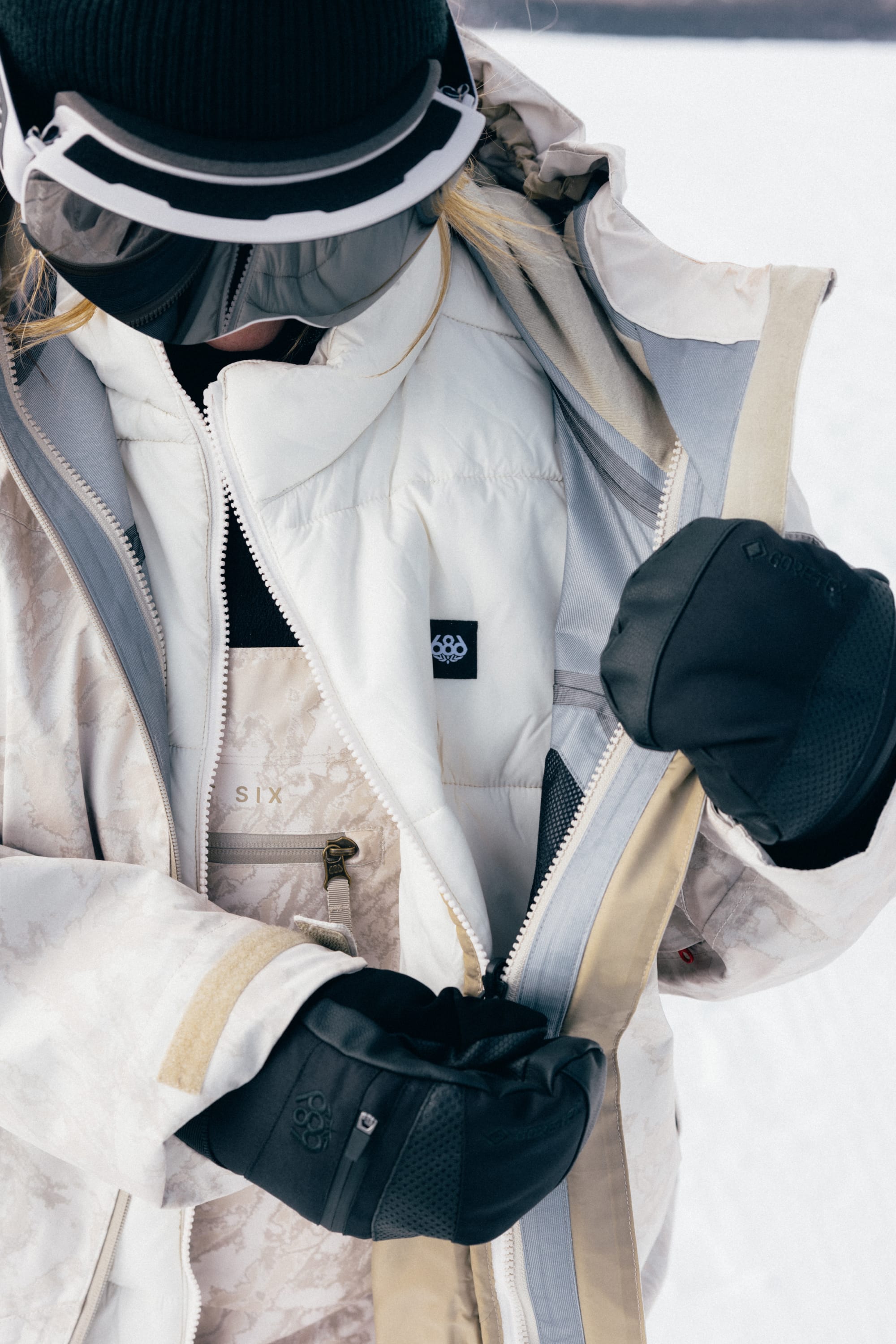 A person wearing the 686 Women’s SMARTY® 3-in-1 Framework™ 3L Jacket and black gloves zips up their coat. They pair it with a black beanie and ski goggles, standing outdoors in a snowy setting.