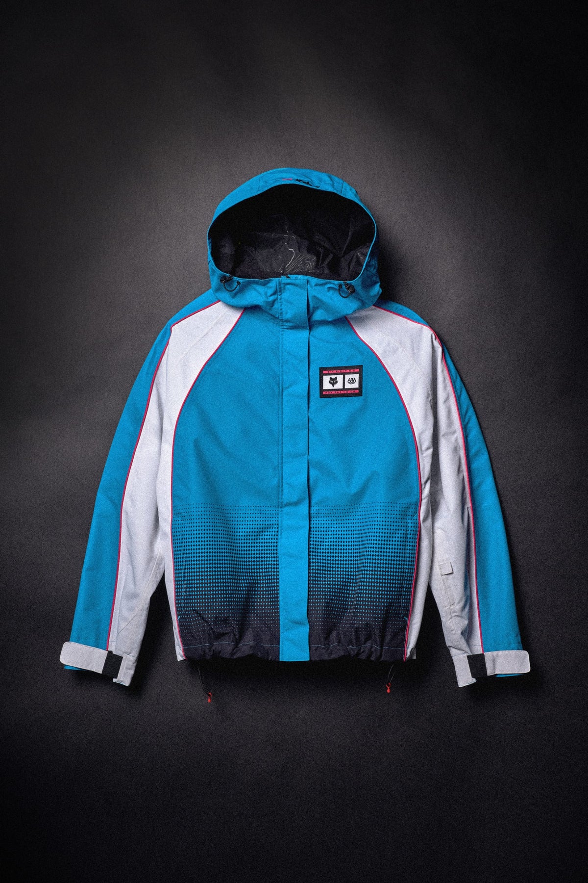 A blue and white 686 Women’s Fox Racing 97 Shell Jacket featuring a hood, a black gradient pattern at the hem, and a Fox Racing logo patch on the chest.