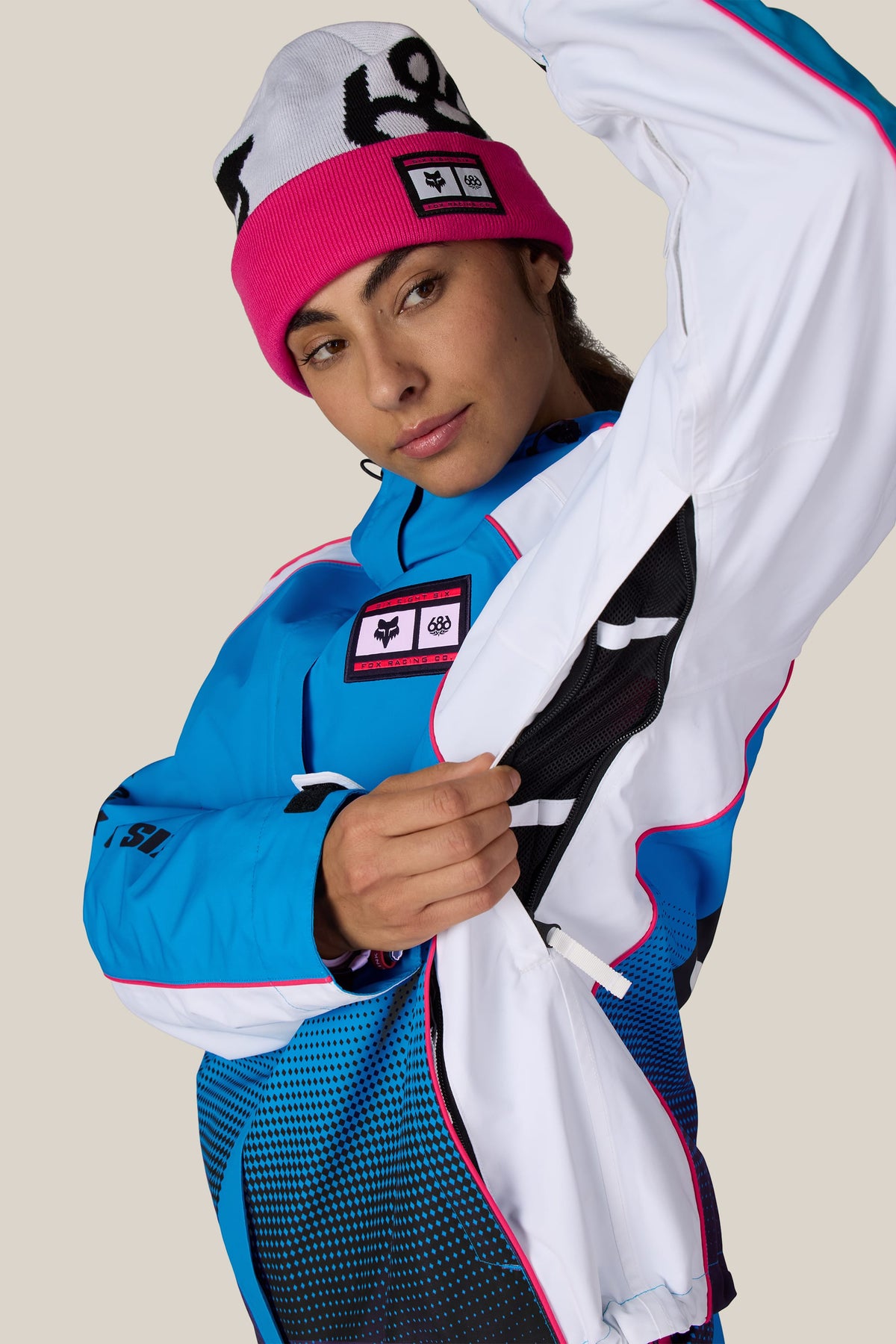 A person wearing a pink and white beanie and a 686 Women’s Fox Racing 97 Shell Jacket, demonstrating the side ventilation zipper.