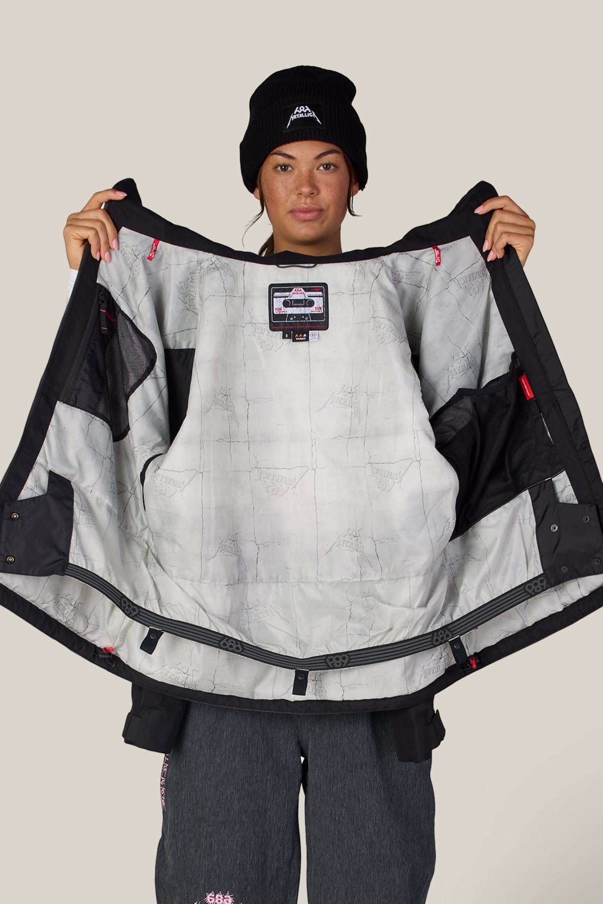 A person holds open a black 686 Women’s Metallica Burning Skull Insulated Jacket to reveal a light gray inner lining printed with a subtle grid pattern and a patch featuring a cassette tape graphic.