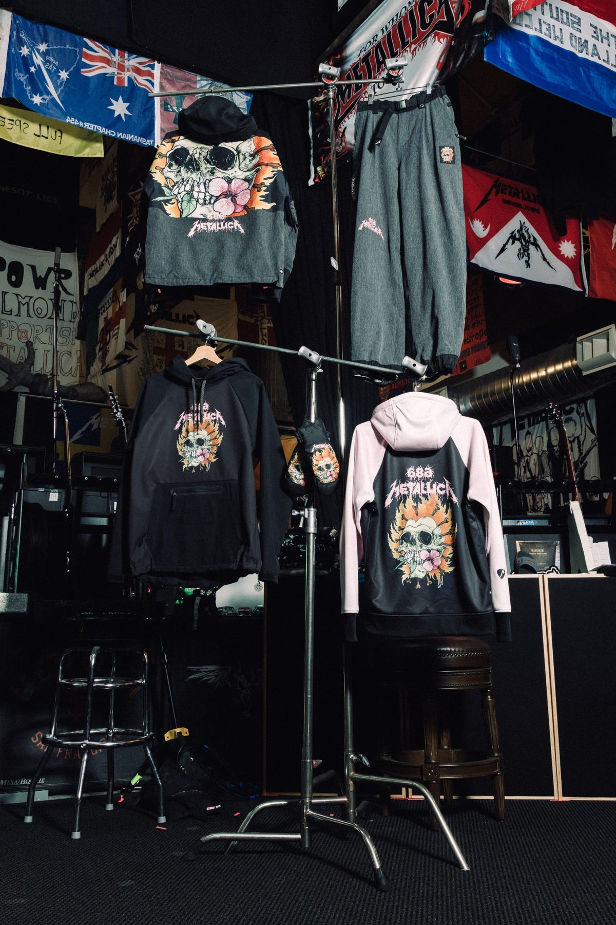 Two 686 Women’s Metallica Burning Skull Insulated Jackets and a pair of pants featuring a graphic of a flaming skull with flowers hang on a clothing rack in a music-themed room.