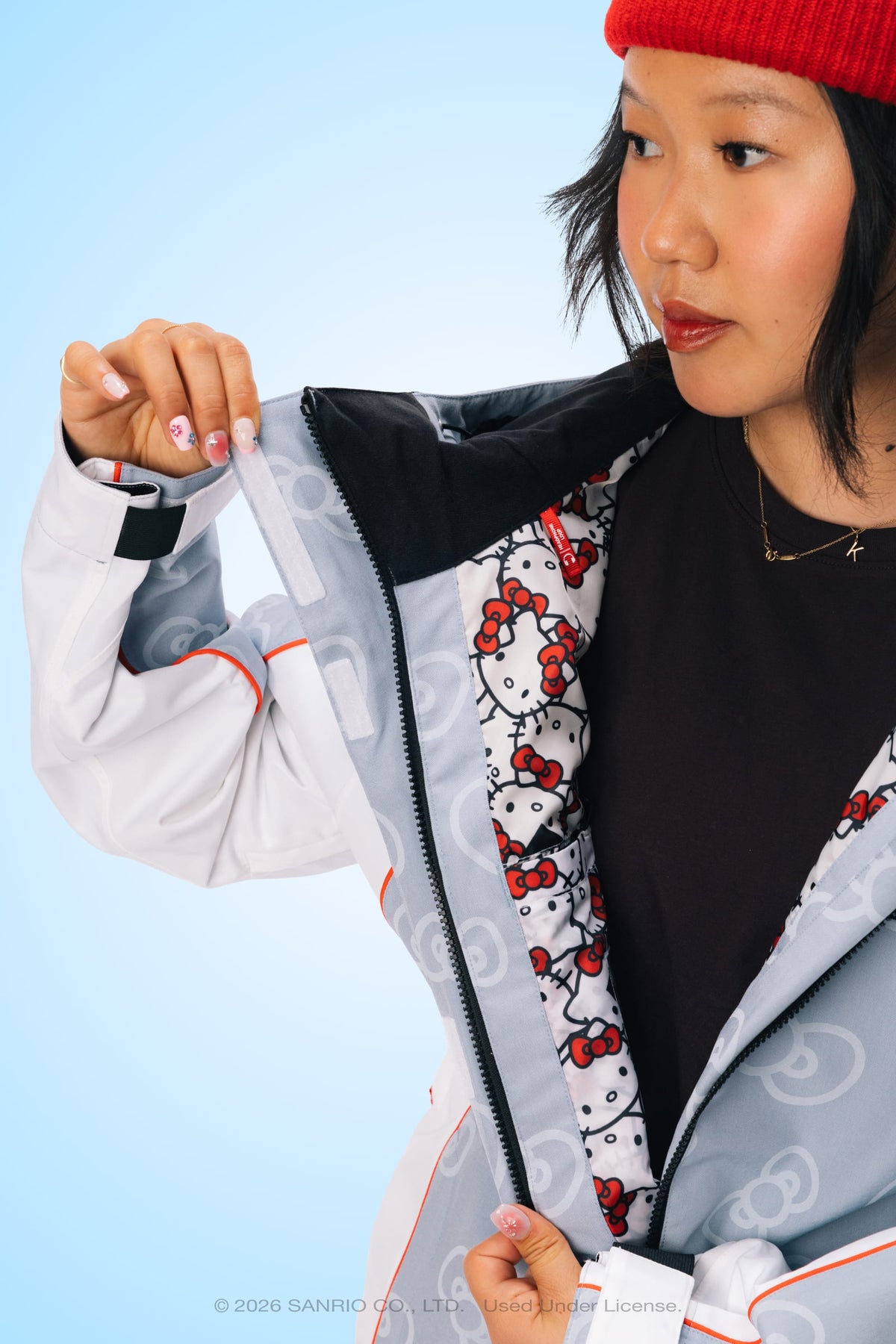 A woman in a red beanie opens her 686 Women’s Hello Kitty® Lil Bow Shell Jacket in light gray, revealing the white Hello Kitty patterned lining. She has painted nails, wears a black shirt and gold necklace, with a blue background behind her.