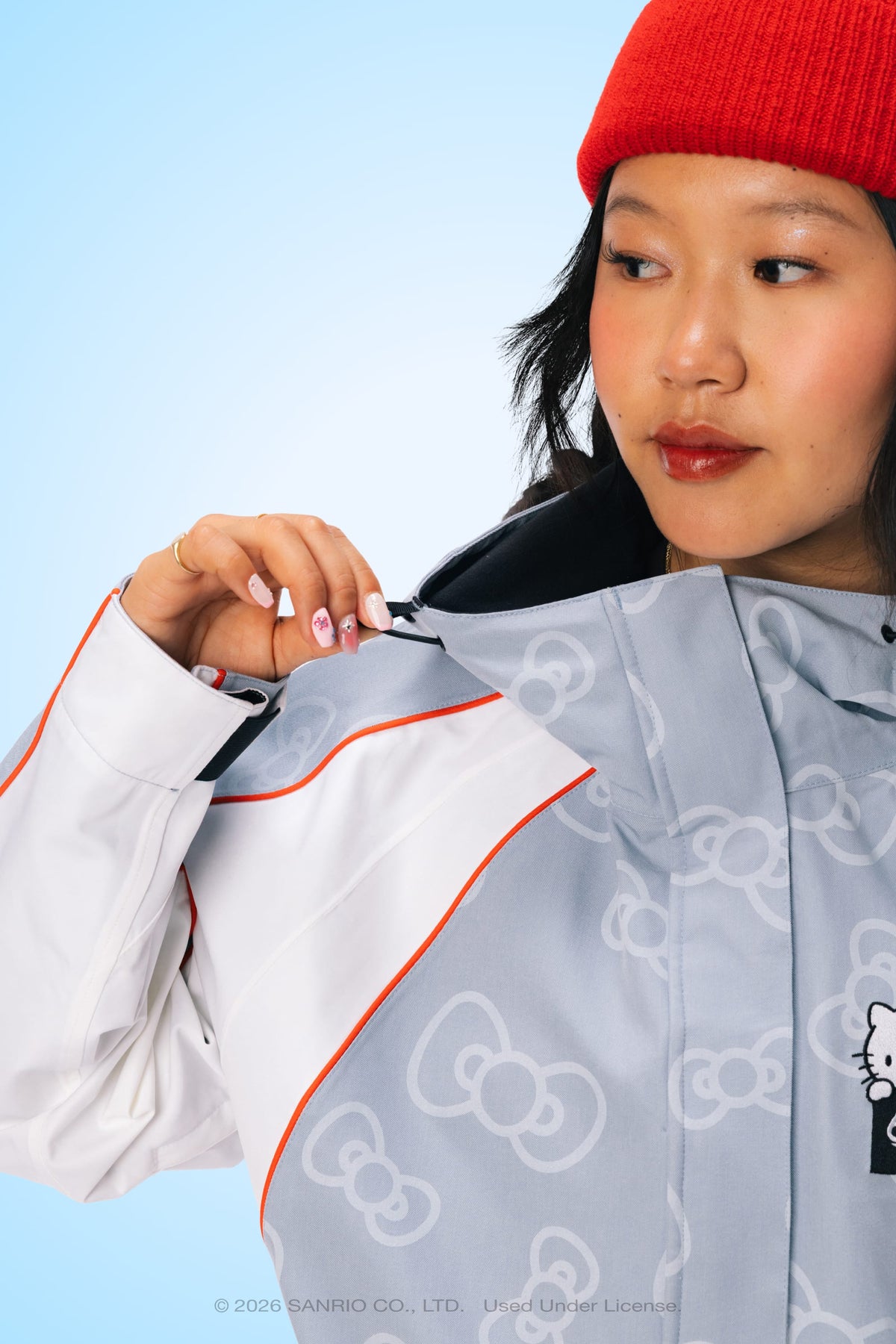 A woman wearing the 686 Women’s Hello Kitty® Lil Bow Shell Jacket by 686, partially unzipped to reveal her fingernail art, and sporting a red beanie, looks to the side against a light blue background.
