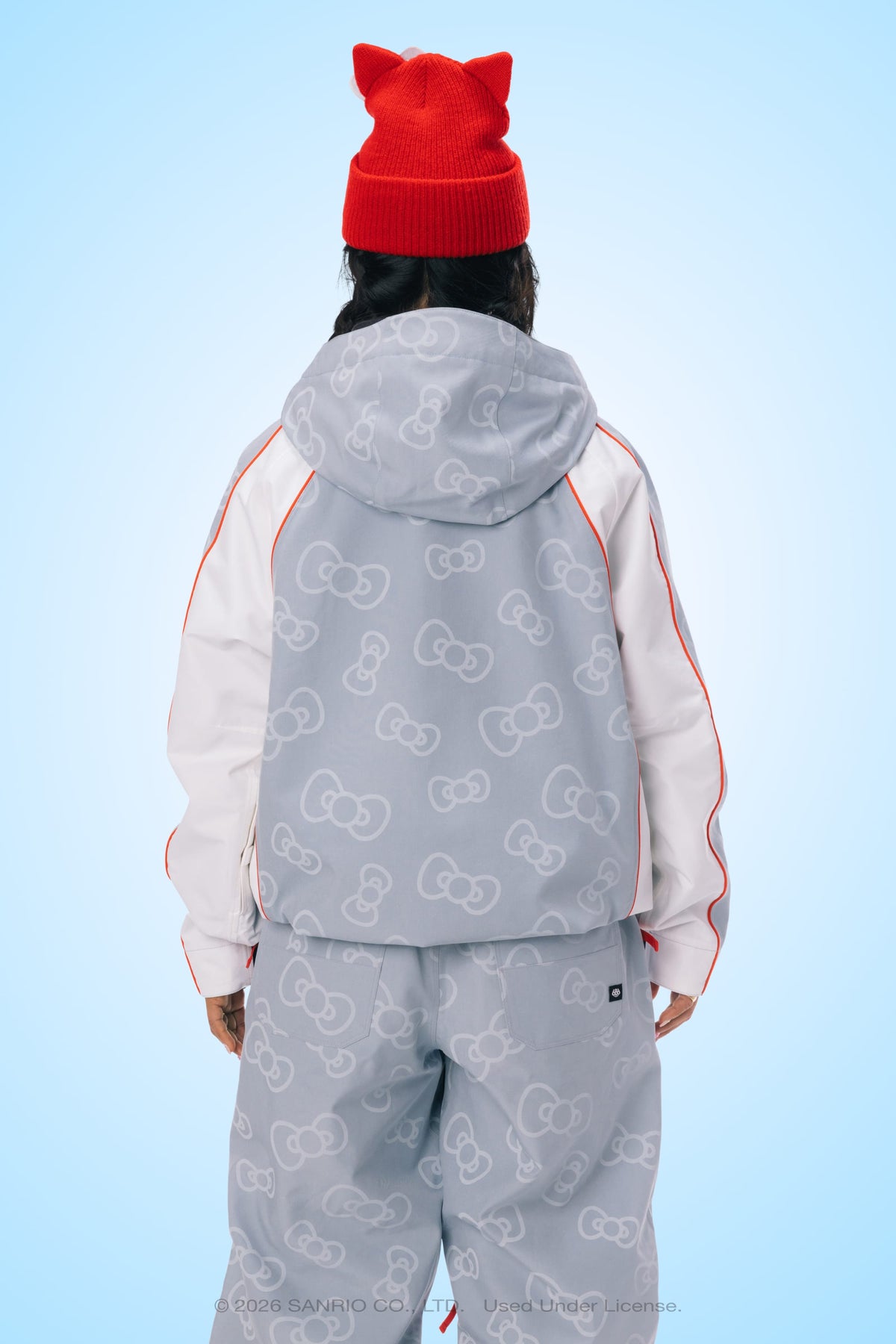 A person facing away, wearing a red beanie hat with cat-like ears and the 686 Women’s Hello Kitty® Lil Bow Shell Jacket by 686, featuring a light gray color with a Hello Kitty bow pattern, along with matching pants, set against a light blue background.