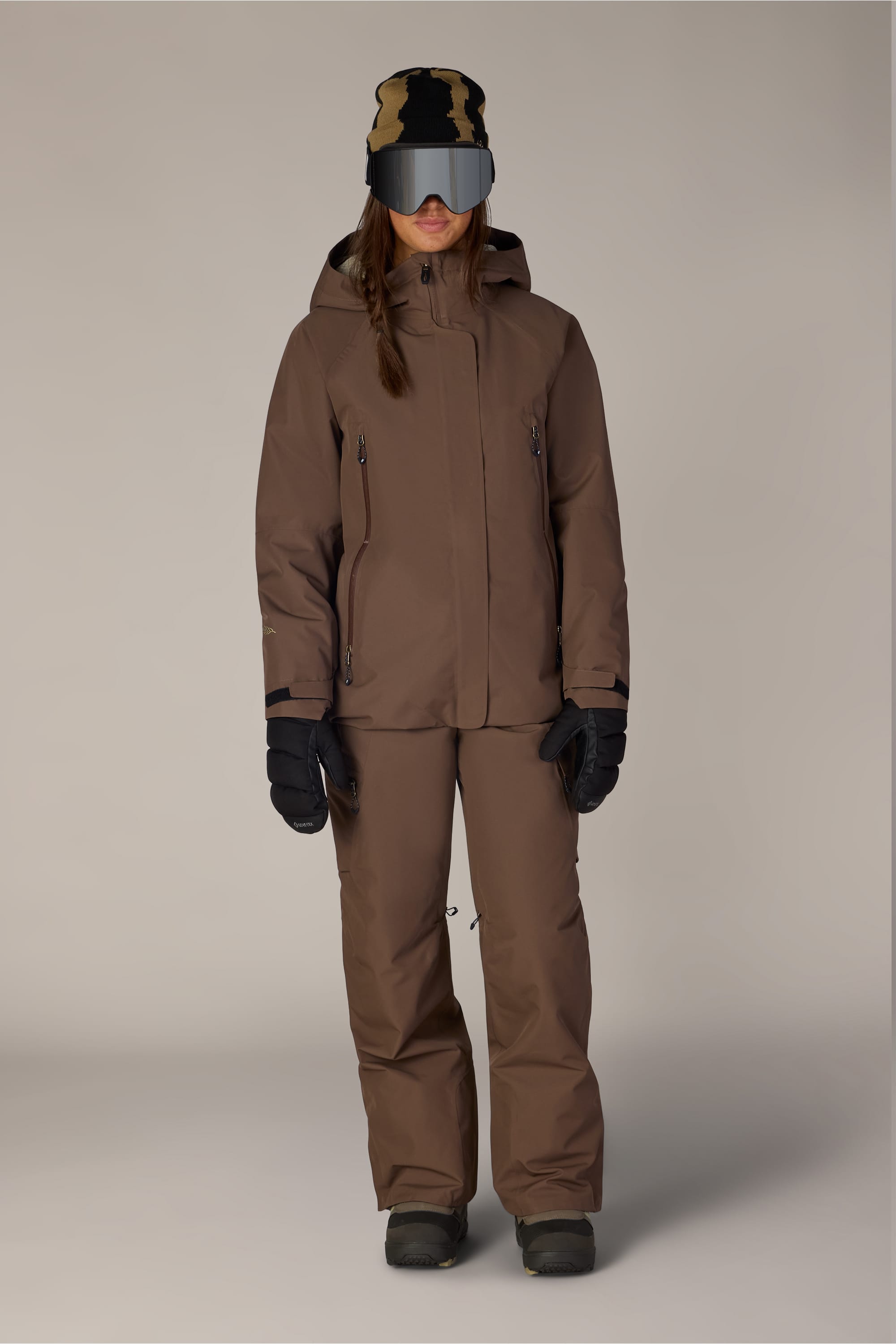 A person wearing a brown winter jacket and the 686 Women’s Geode Thermagraph® Pant, black gloves, dark boots, a black and tan hat, and large reflective ski goggles stands against a plain light background.