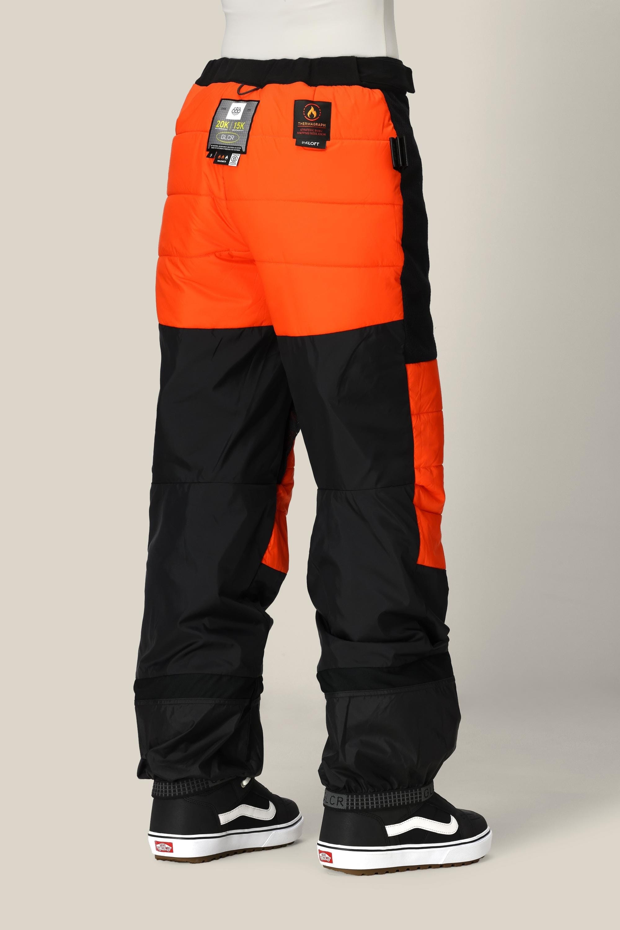 A person is standing and facing away, wearing 686 Women’s Geode Thermagraph® Pant in black and orange with tags attached. They are also wearing black and white slip-on sneakers, visible from behind. The background is a plain light color.