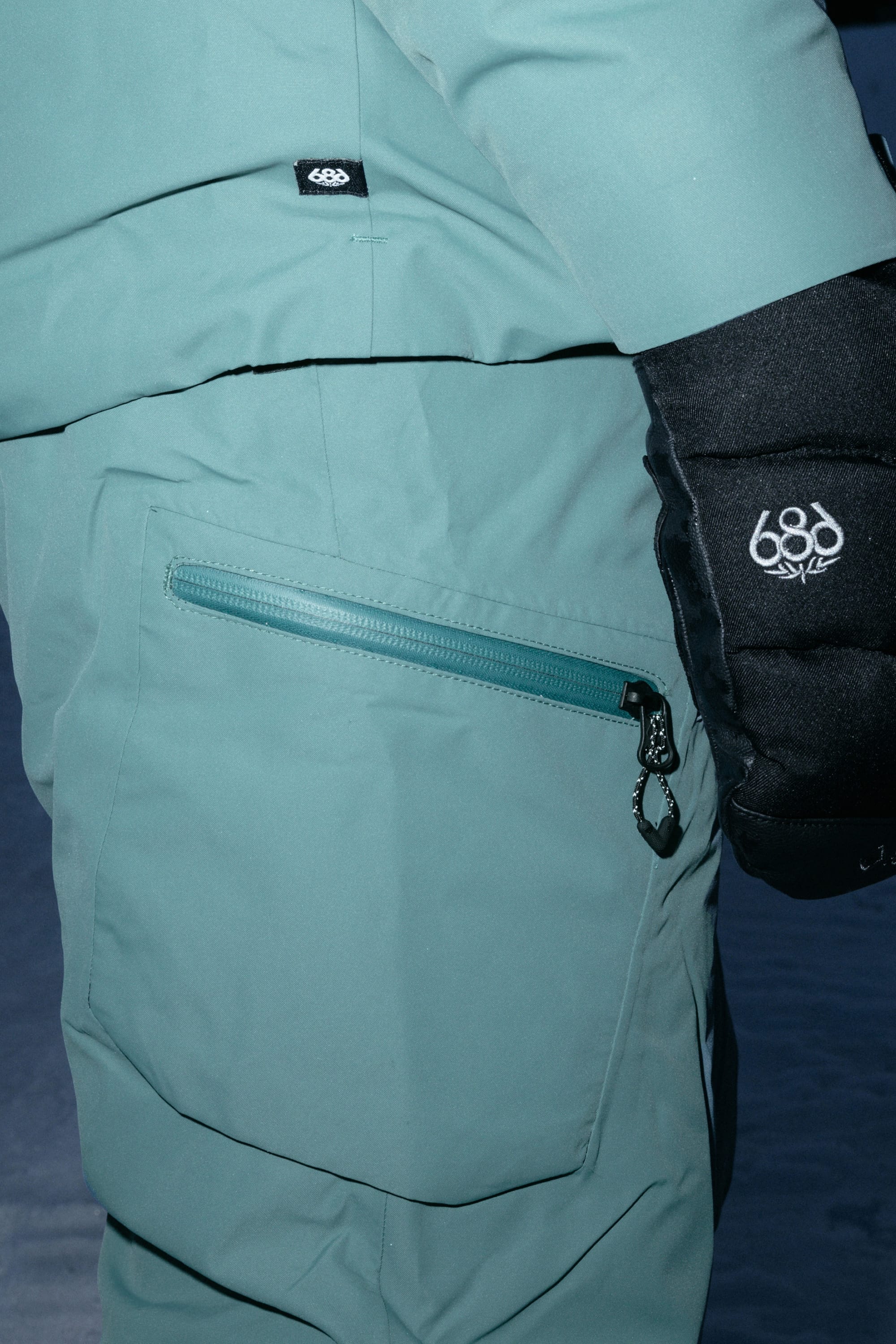 Close-up of a person standing on snow wearing the 686 Women’s Geode Thermagraph® Pant in teal, made from durable polyester with a zippered pocket and sporting a black glove—both featuring the 686 logo.