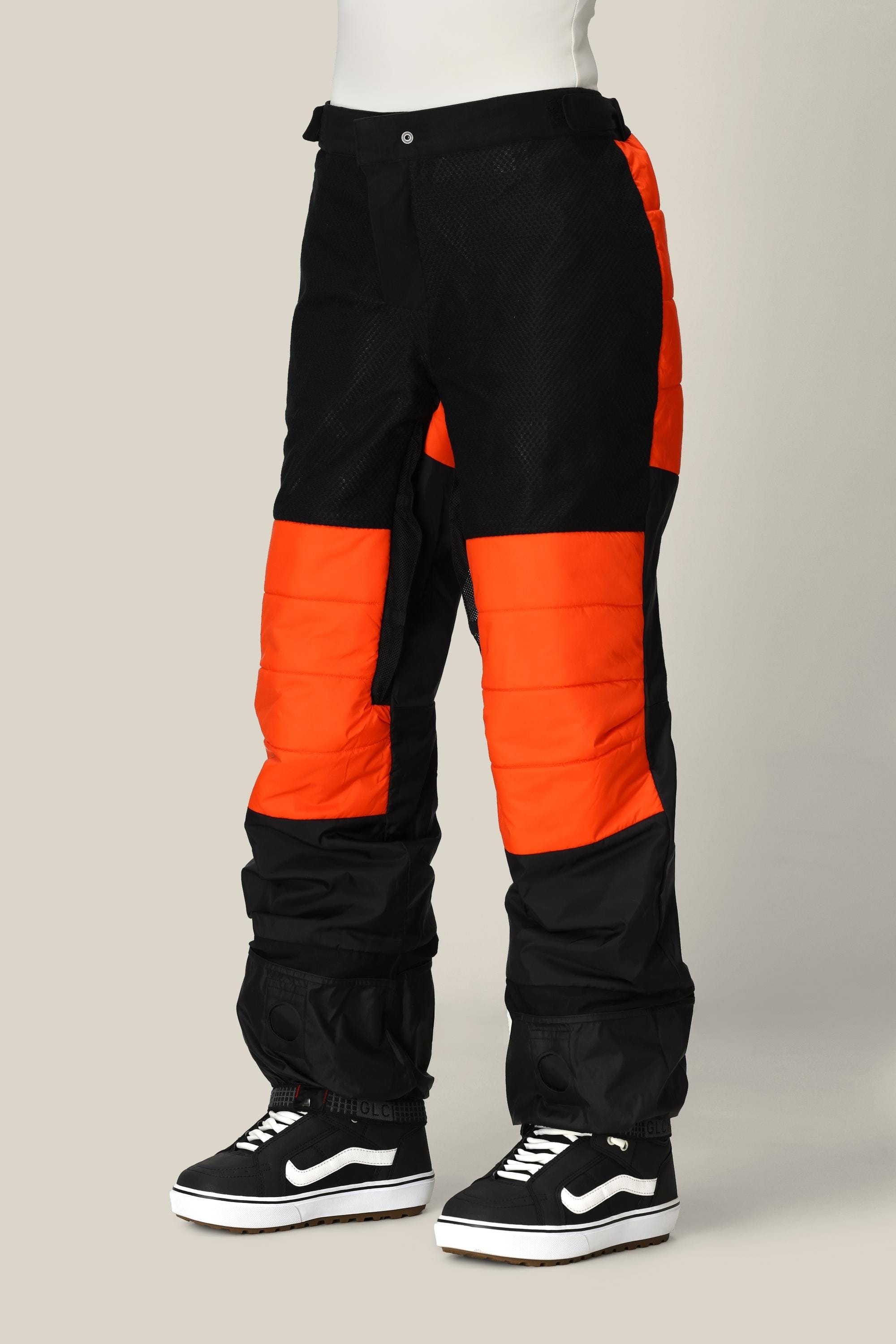 A person is wearing the 686 Women’s Geode Thermagraph® Pant by 686—these women’s waterproof pants feature a striking black and bright orange colorblock design, padded sections, a loose fit, and are crafted from durable waterproof polyester—paired with black Vans sneakers. The upper body is not visible, and the background is a plain neutral color.