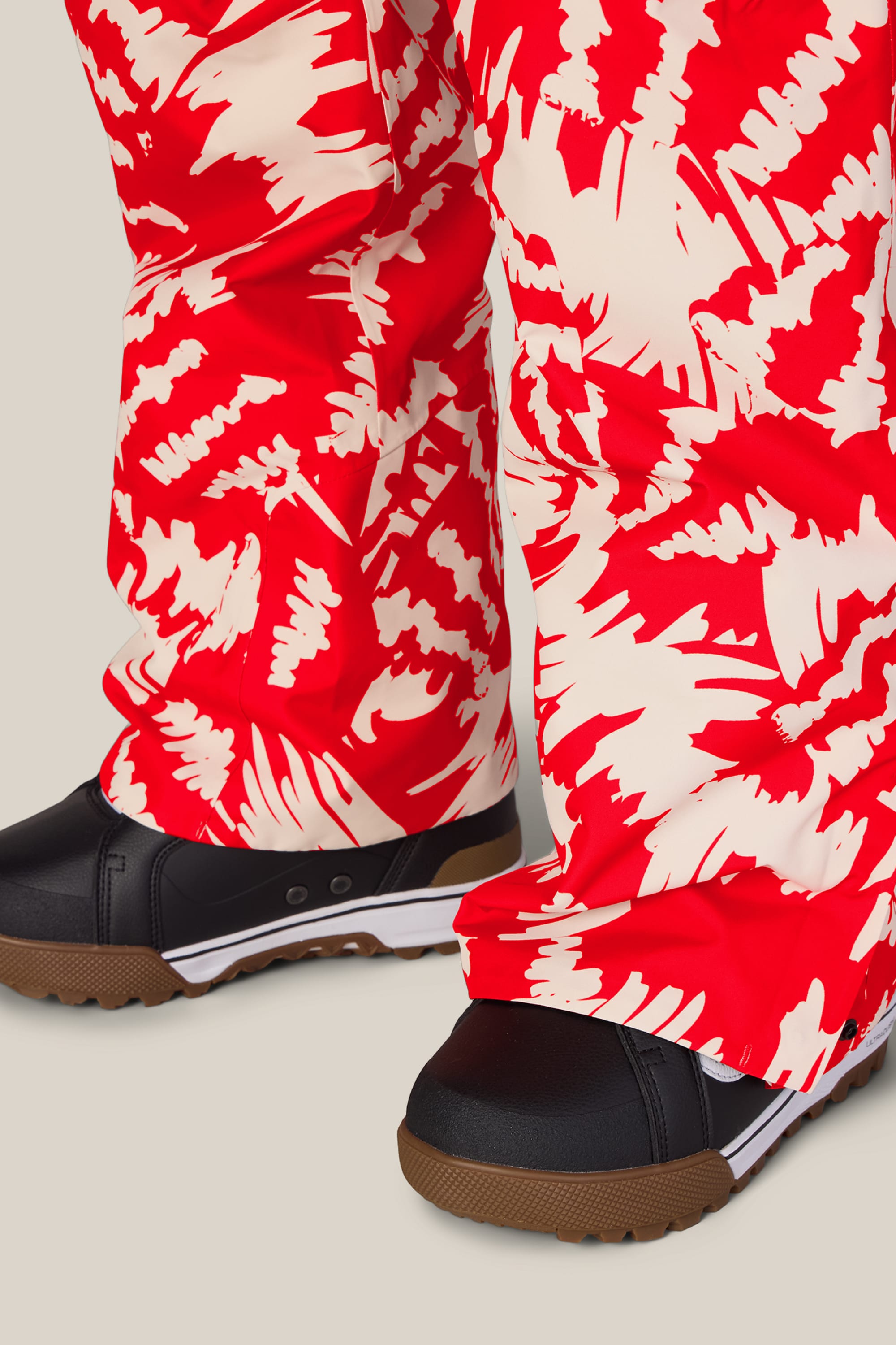 Close-up of a person wearing the 686 Women’s Geode Thermagraph® Pant by 686, featuring a red and white pattern, paired with black shoes with brown soles, standing on a light background.