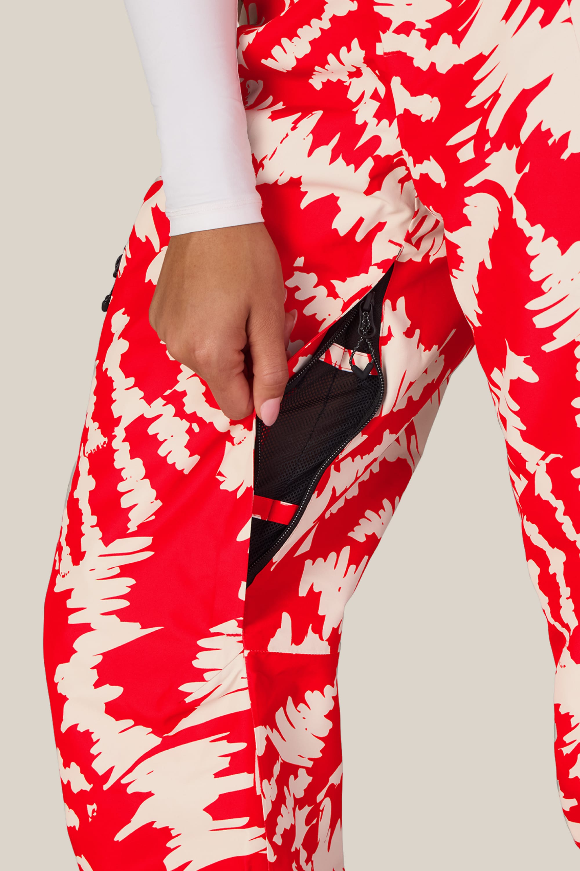 A woman wearing the 686 Women’s Geode Thermagraph® Pant in a striking red and white pattern stands with her hand pulling open the zipper of a mesh pocket, revealing the partially open compartment. These stylish waterproof pants from 686 seamlessly blend fashion and function, ensuring you stay dry and comfortable.