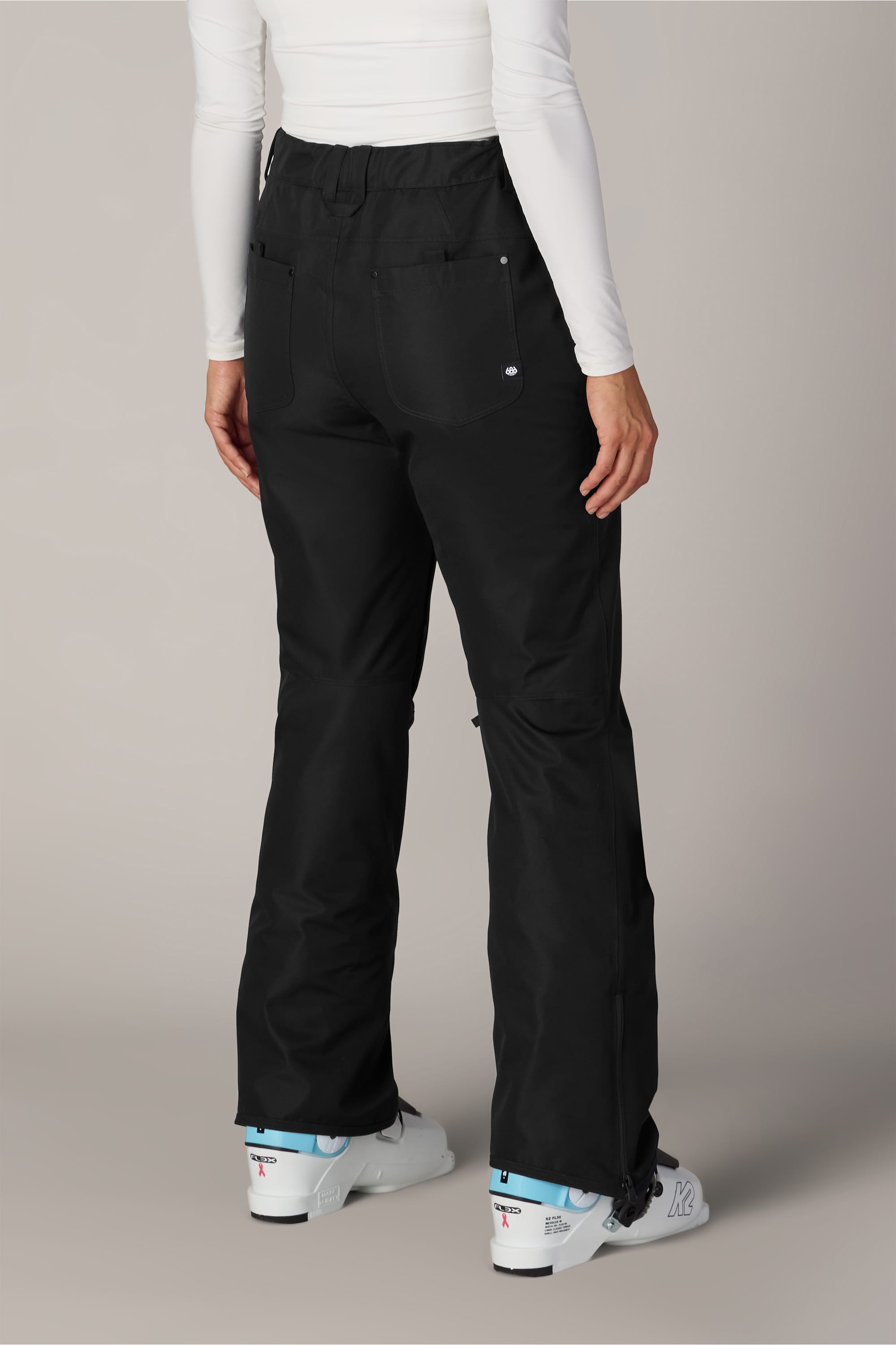 A person is shown from the back, standing on a plain, light-colored background, wearing the 686 Women’s Slim Line Pant in black polyester waterproof fabric, paired with a white long-sleeve top and white ski boots.