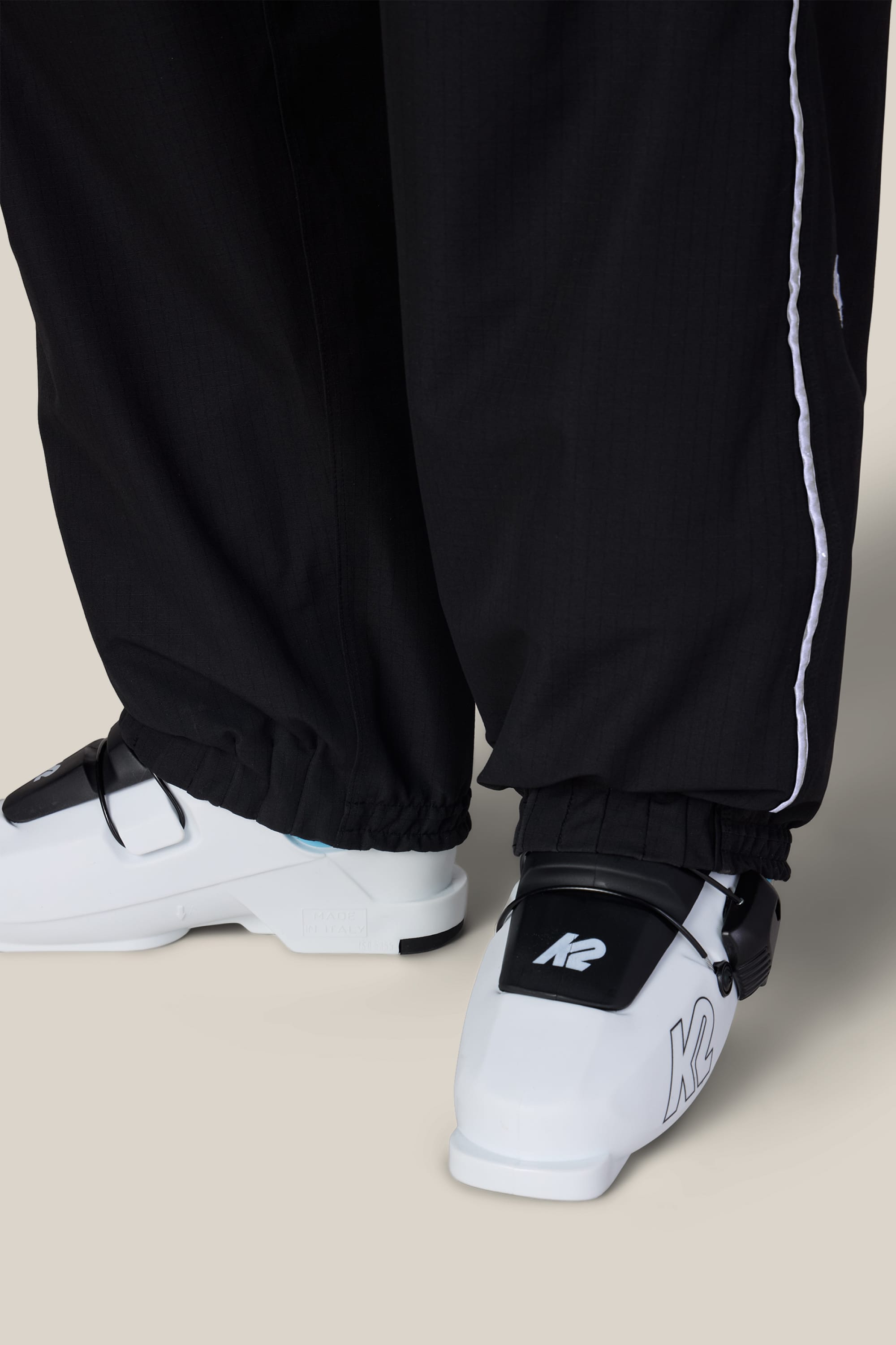 A woman wears 686 Women’s Hellkat™ Shell Pants with elastic cuffs and white ski boots featuring the K2 logo, posed against a light background.