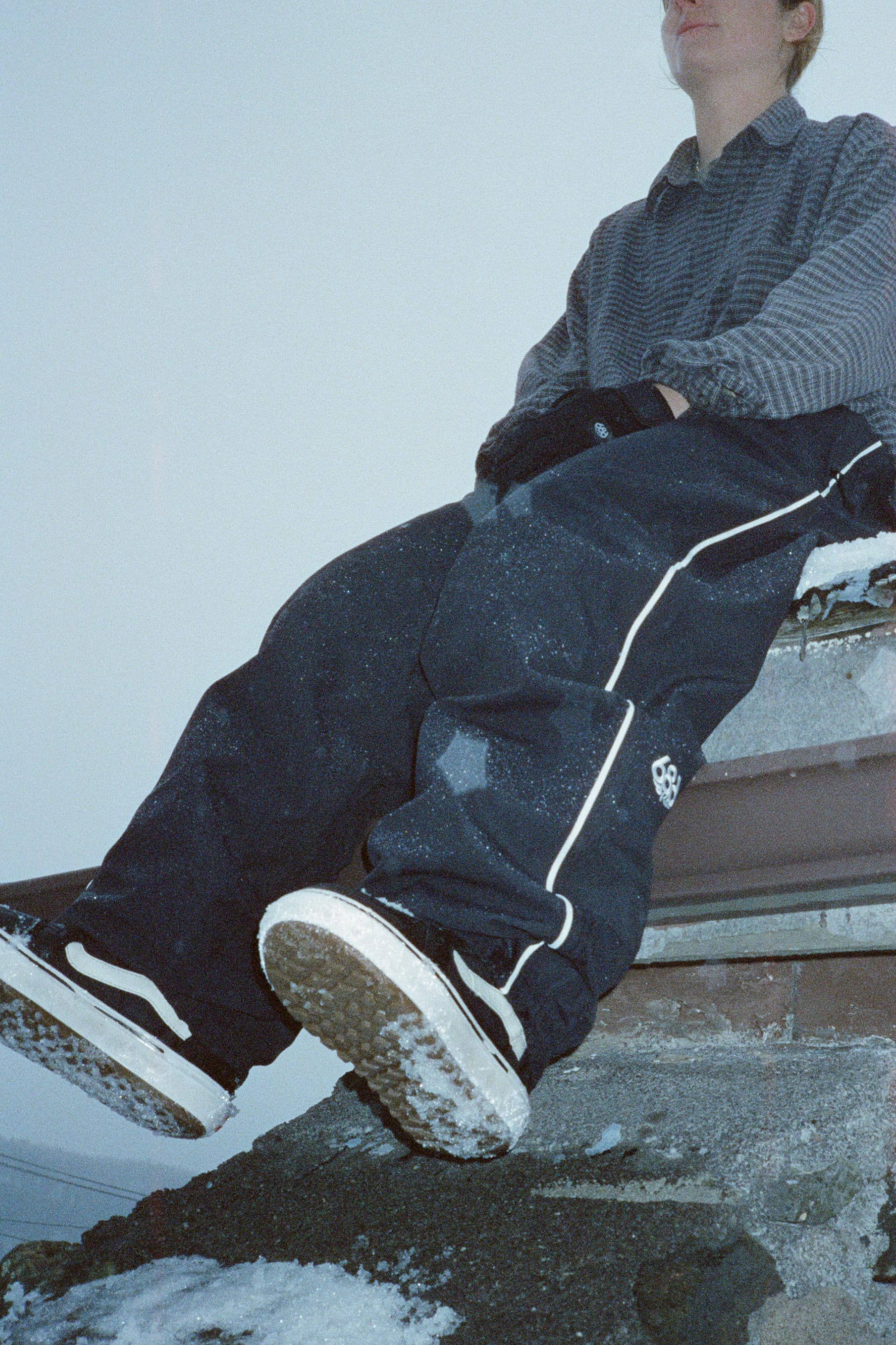 Wearing a checkered shirt and 686 Women’s Hellkat™ Shell Pants, a person sits on a snowy rooftop. The photo highlights their legs and shoes, with snow scattered on the ground and their clothing.