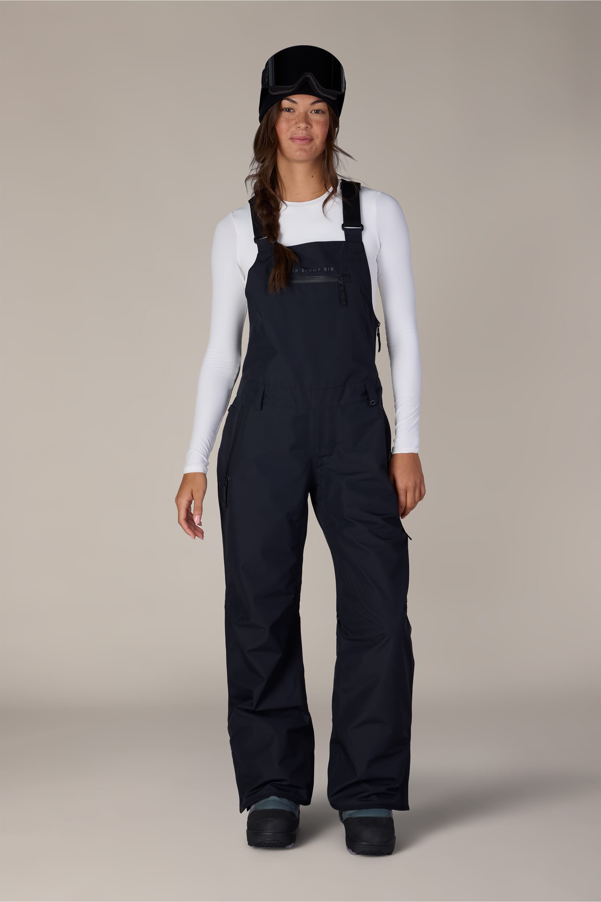 A person wearing a white long-sleeve shirt, the 686 Women’s Regalia™ Bib snow pants in black, black snow boots, and a black ski helmet with goggles stands against a plain background.