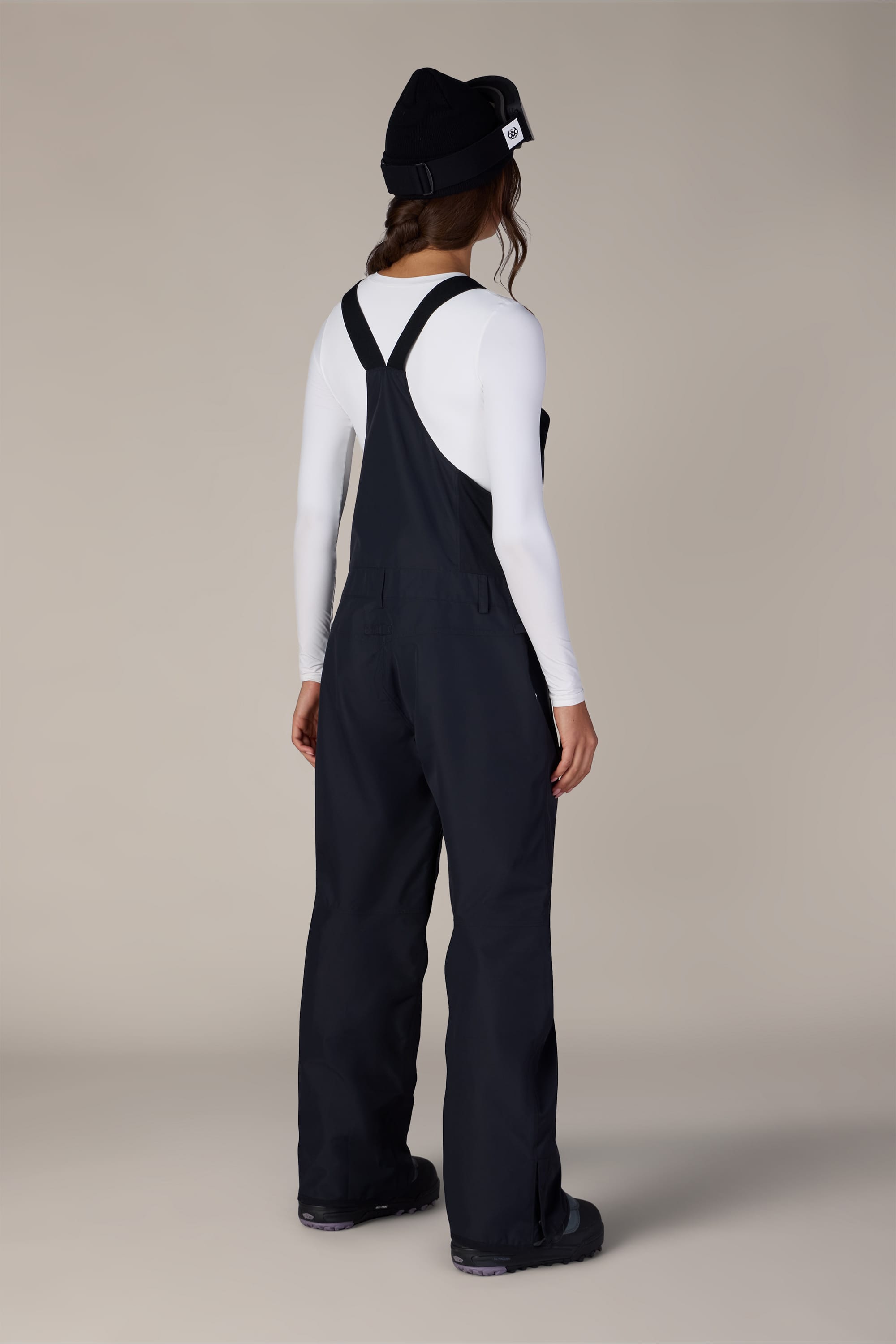 A person stands facing away, wearing a black beanie, a white long-sleeve shirt, and the 686 Women’s Regalia™ Bib by 686—black polyester bib overalls with wide straps. The background is plain and neutral.
