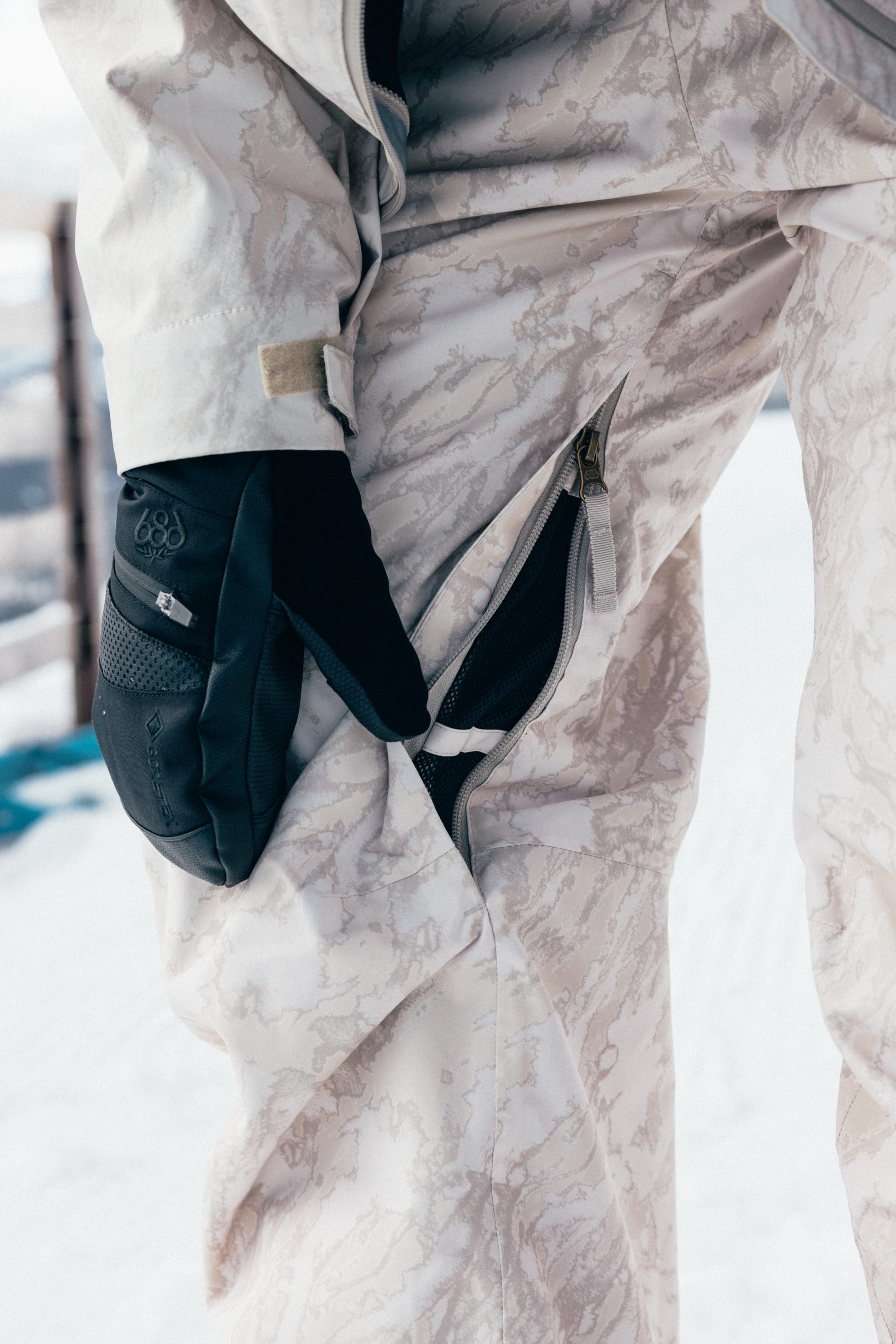 A person wearing the 686 Women’s Regalia™ Bib snow pants and black gloves is unzipping a vent on the side of their pants, with a snowy background visible.
