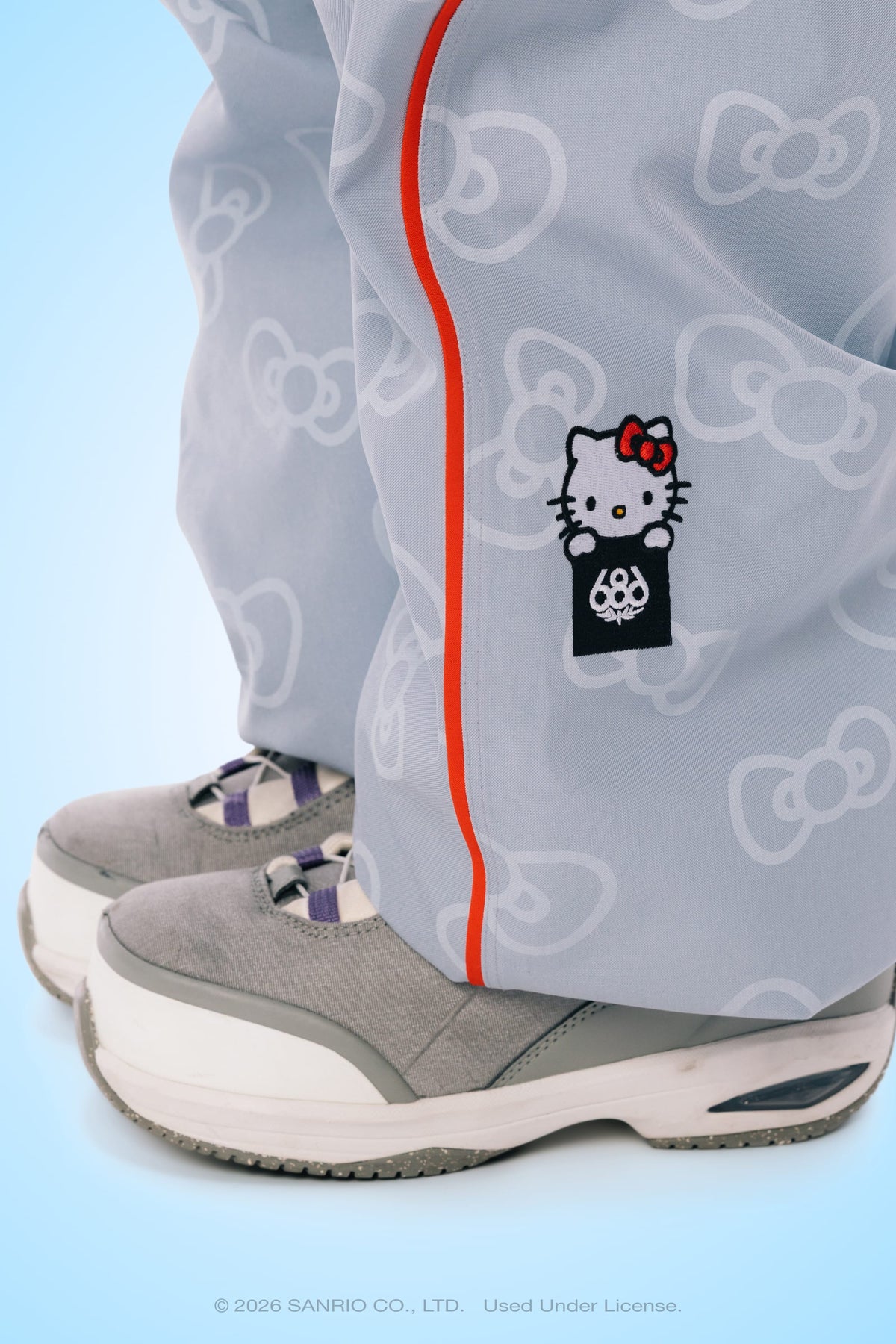 Close-up of a person wearing the 686 Women’s Hello Kitty® Lil Bow Shell Pant in light gray, featuring a Hello Kitty patch and red piping, paired with gray and white sneakers. The background is light blue.