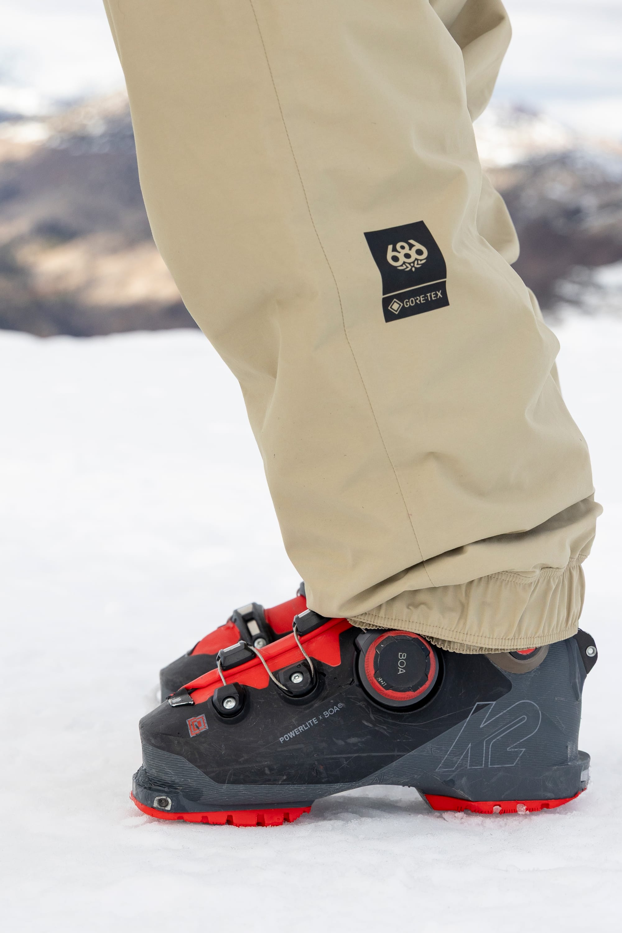 Wearing the 686 Women’s GORE-TEX® Outline™ Pant, a person stands on snow in front of a blurred mountain backdrop, paired with black-and-red ski boots.