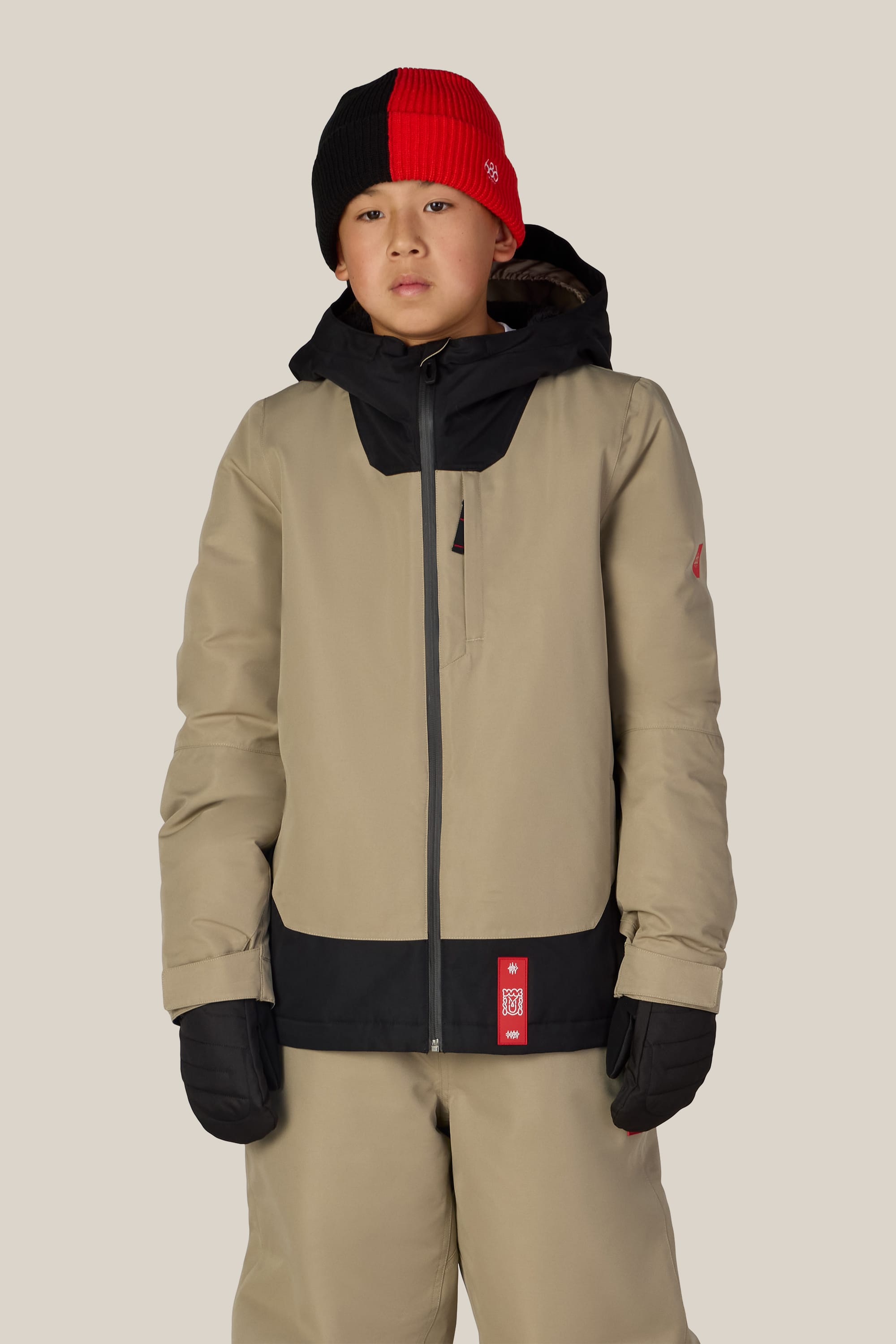 A young person wearing the 686 Boys’ Gateway™ Insulated Jacket in tan and black, with matching pants, black gloves, and a red and black beanie stands facing forward against a plain beige background.